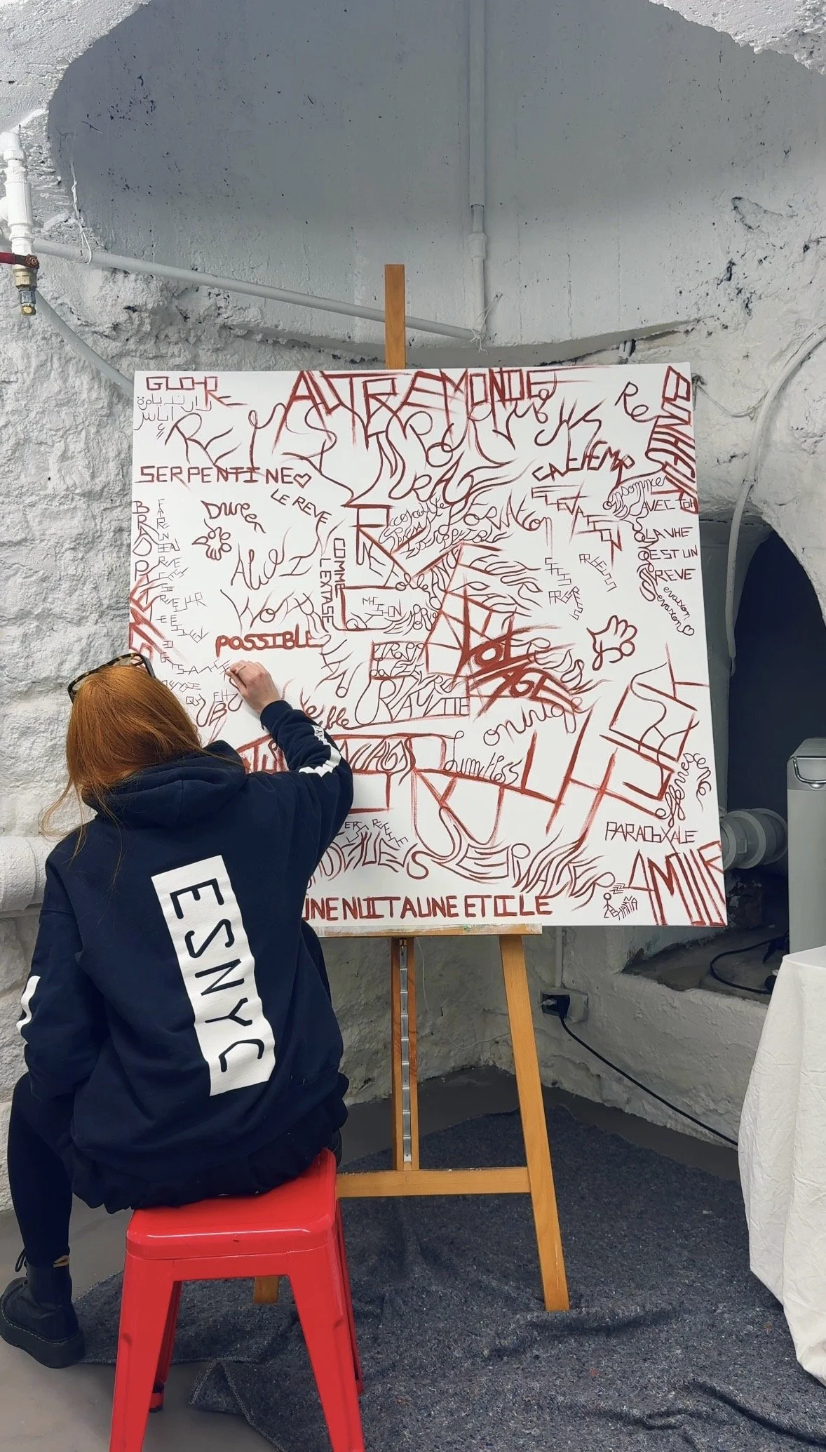 Manon, rousse, assise, peinant une toile collaborative fait a partir des mots des visiteurs, thème REVE, fond blanc, dans son atelier dans un concept store.