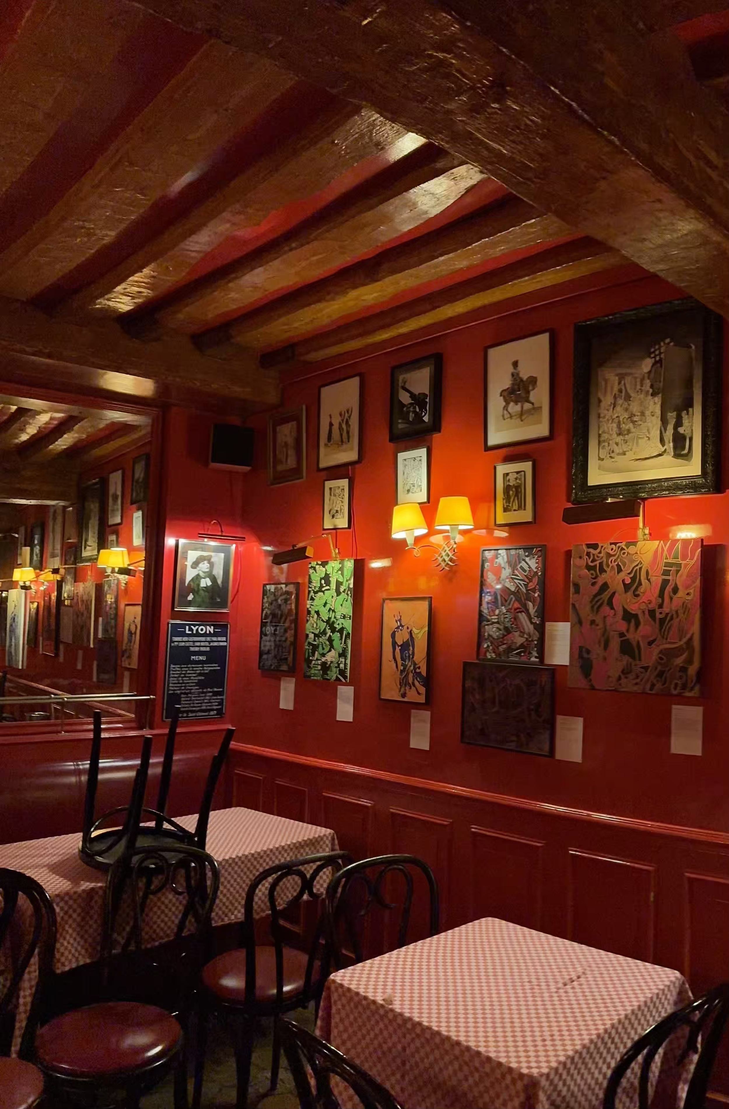 Restaurant chez Castel où sont exposés les oeuvres de Manon Sajous, œuvres d'art accrochées à un mur rouge, avec des tables couvertes de nappes à carreaux rouges et blancs, et des chaises en bois.