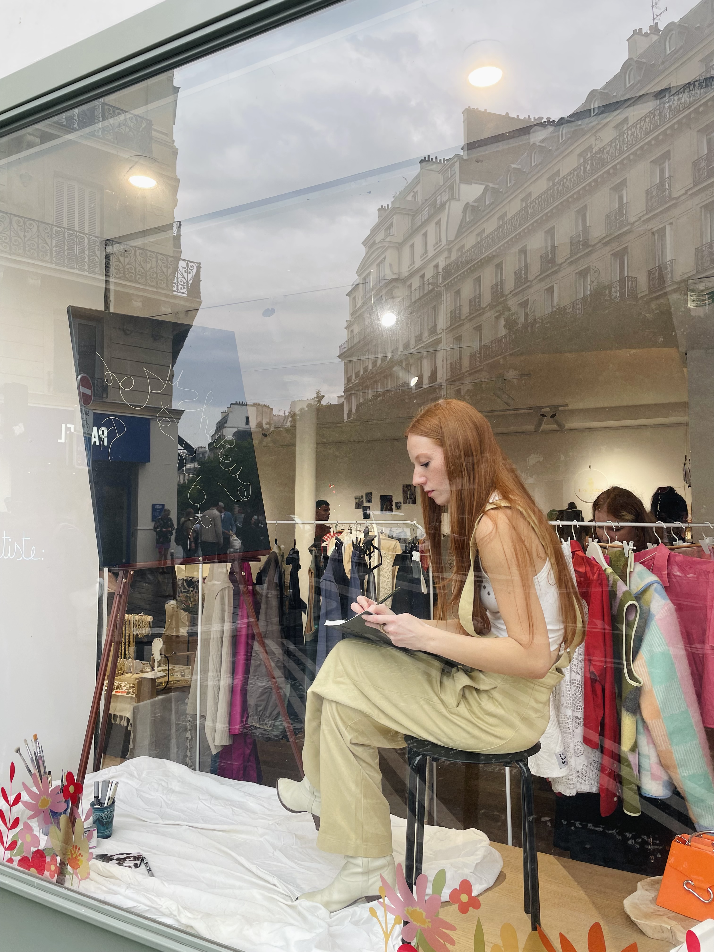 Vitrine d'un pop up avec une jeune femme aux cheveux roux (Manon SAJOUS) assise sur un tabouret, écrivant sur son cahier, les mots qu'elle va écrire sur la toile.
