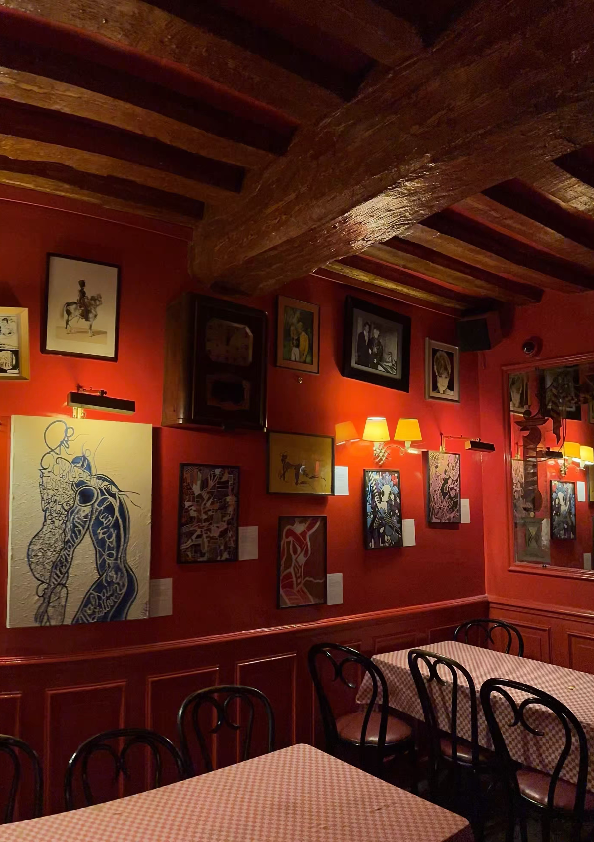 Restaurant chez Castel où sont exposés les oeuvres de Manon Sajous, œuvres d'art accrochées à un mur rouge, avec des tables couvertes de nappes à carreaux rouges et blancs, et des chaises en bois.