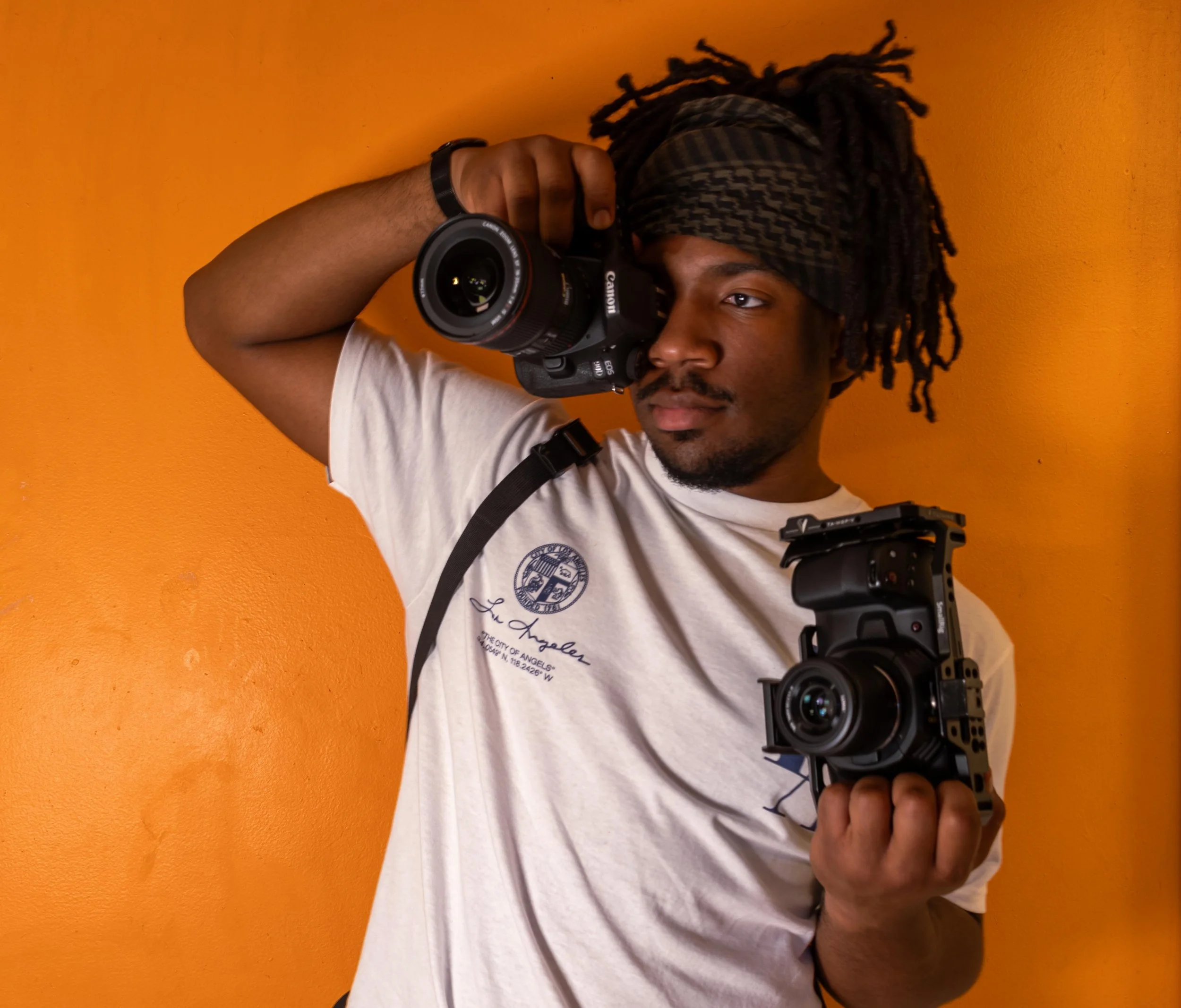 A person with dreadlocks and a plaid headband holding a professional camera, taking a photo against a bright orange wall.