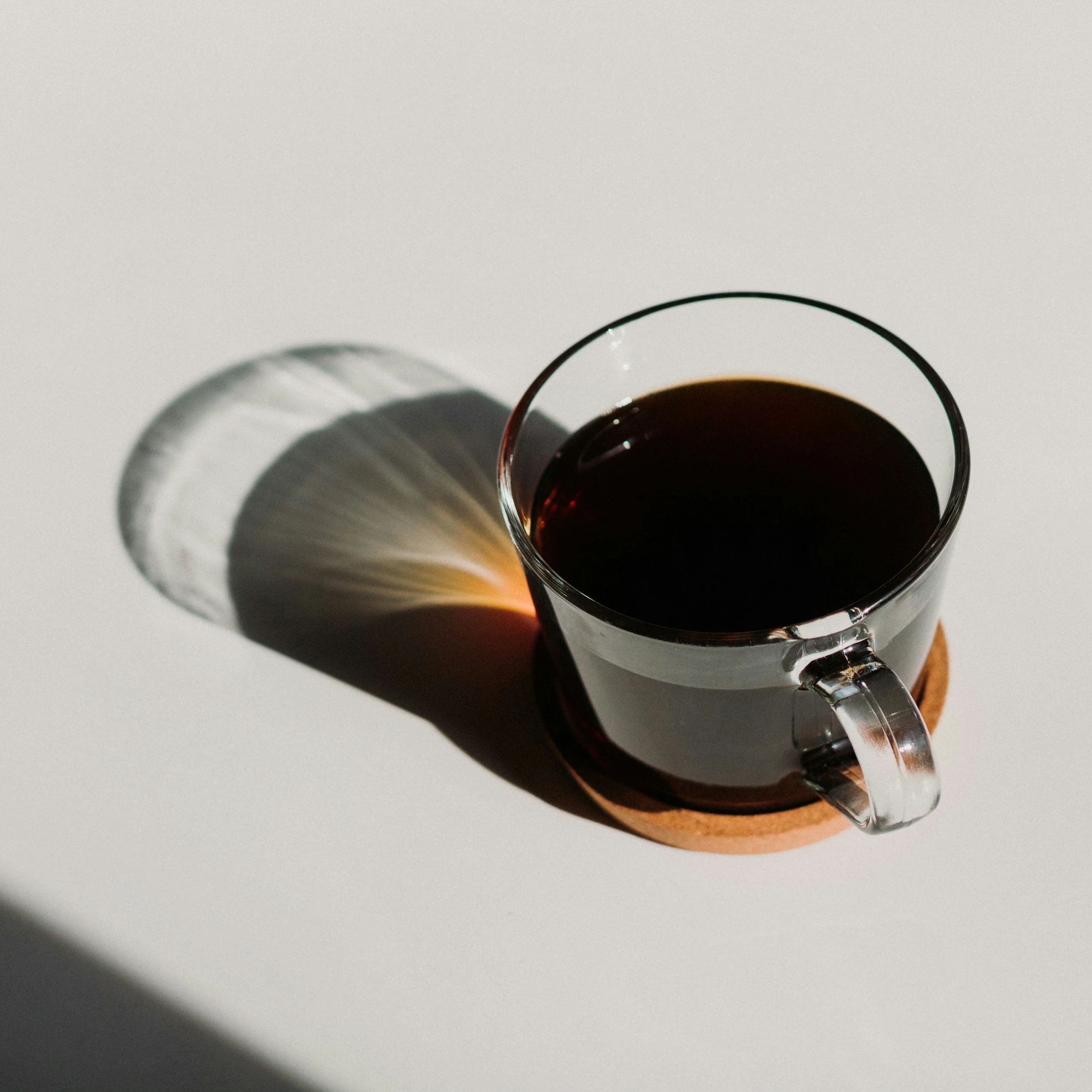 A clear glass cup filled with black coffee placed on a cork coaster. The sunlight creates a colorful shadow and reflection on the white surface below.