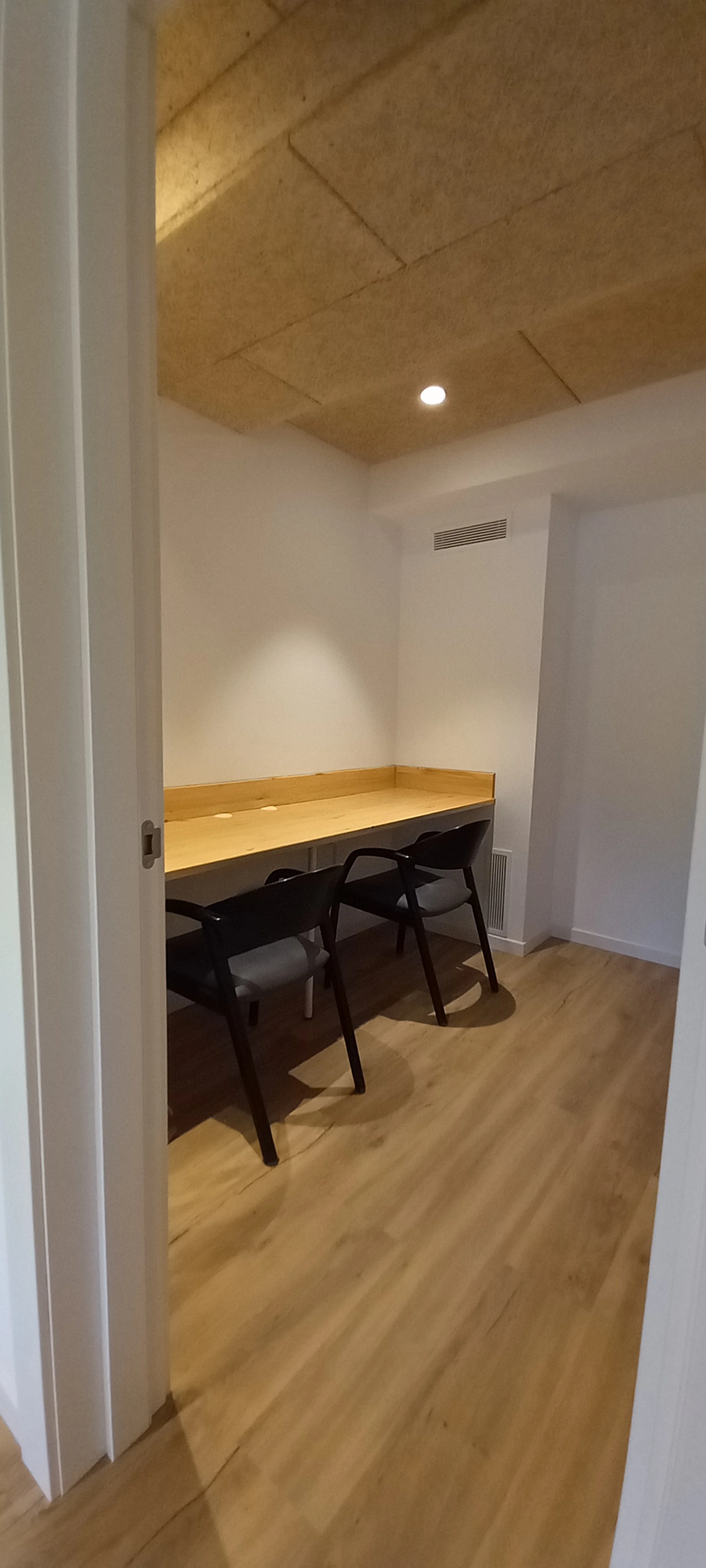 A soundproof booth/small meeting room with a wooden table, two black chairs, white walls, wooden flooring, and a ceiling with acoustic panels and a recessed light.