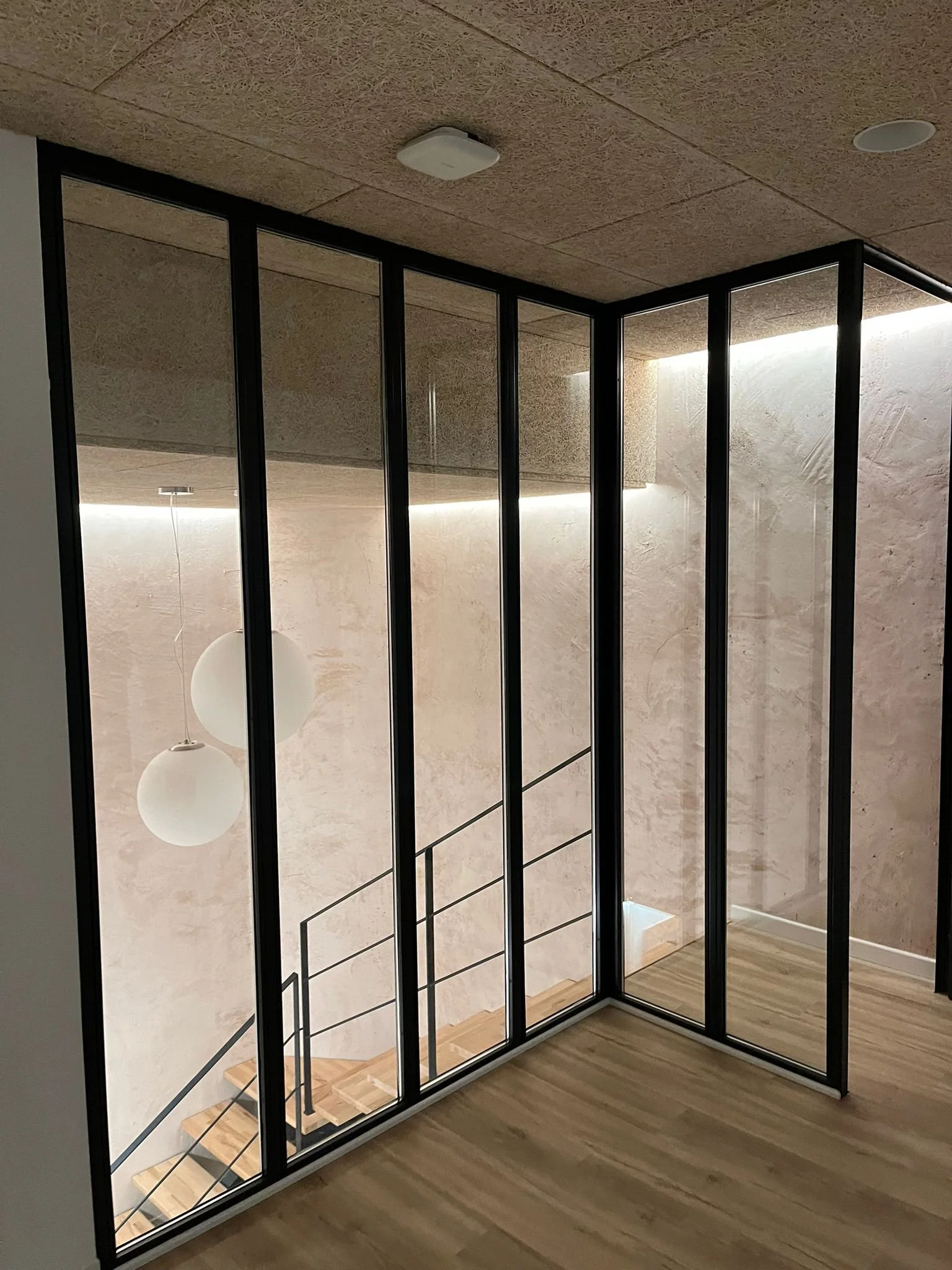 Modern interior with a glass partition, a staircase, two white spherical pendant lights, and a textured wall with soft lighting.