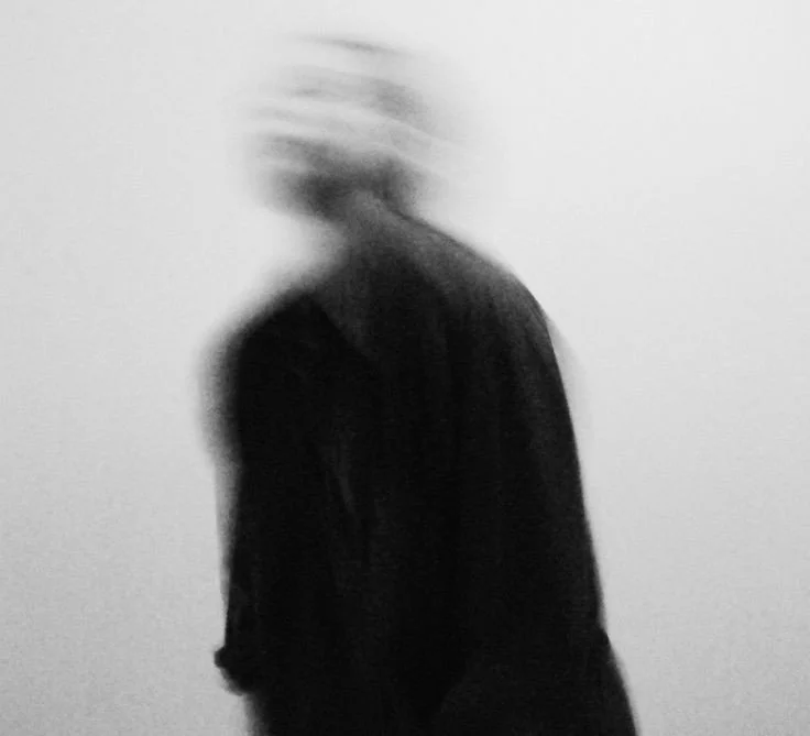 Blurry black and white photo of a person with turned head, wearing a dark jacket, standing against a plain light background.