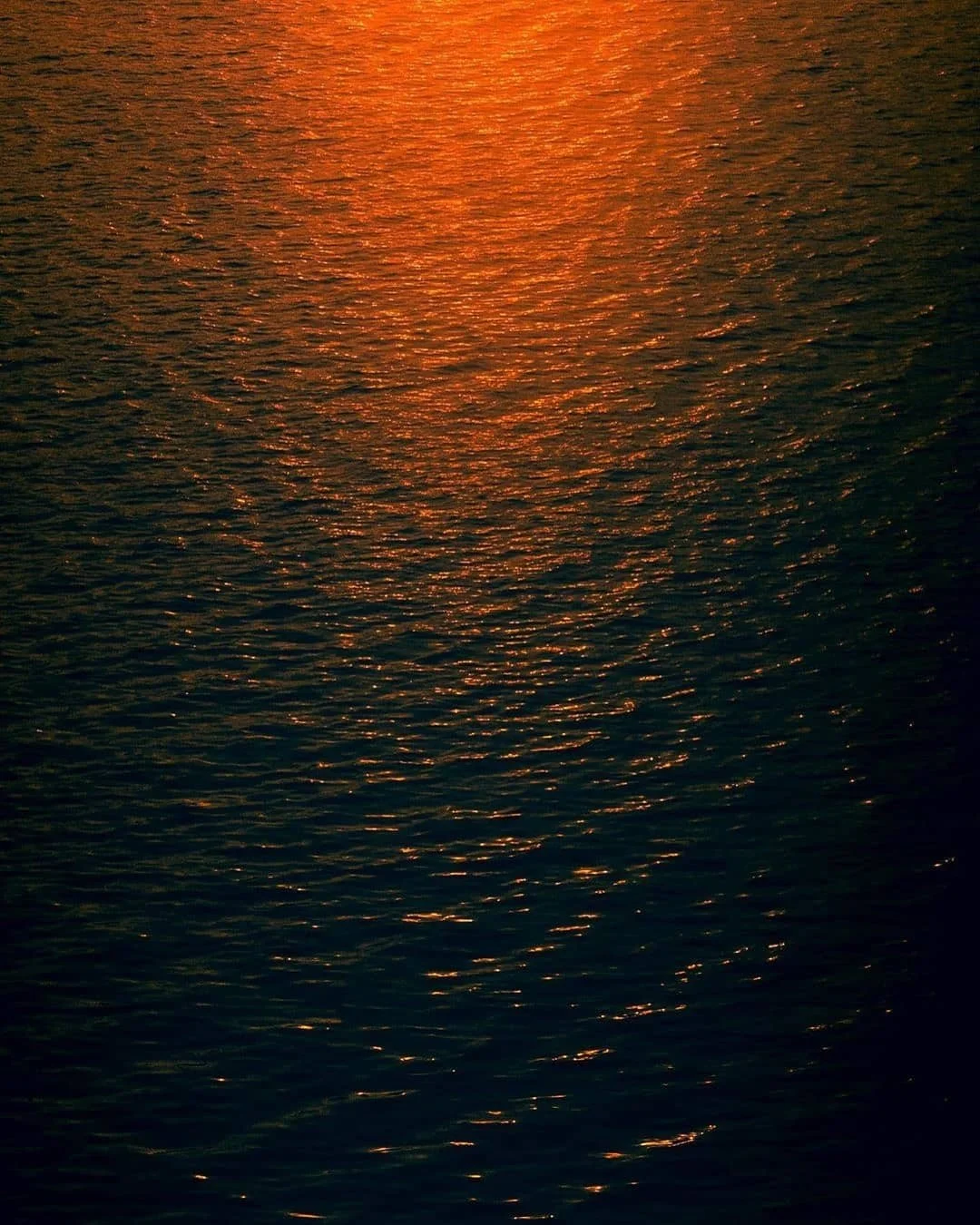 Sunset over a body of water, with reflections creating a shimmering orange glow on the surface.