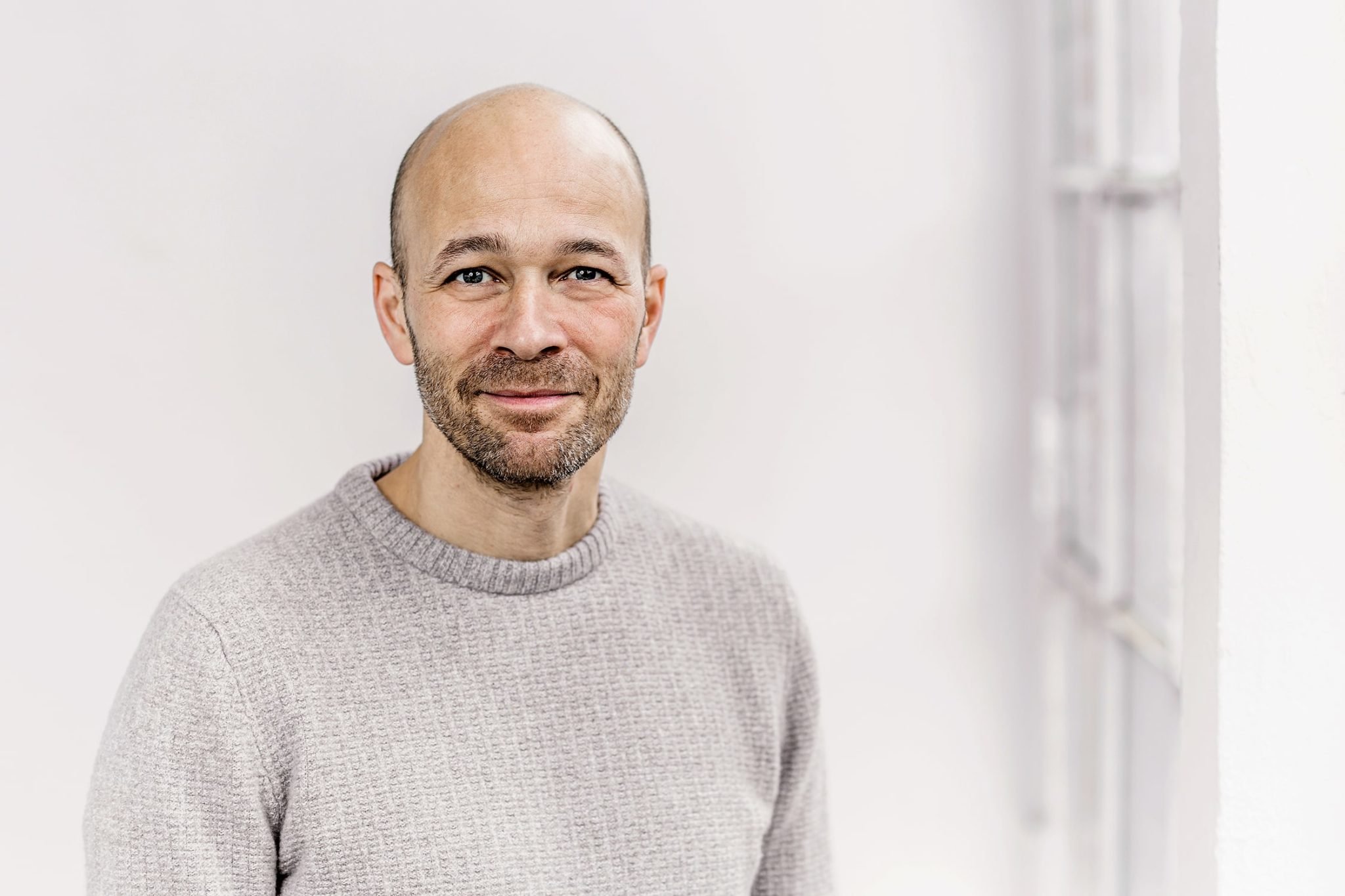 A smiling, bald man with a beard wearing a beige knitted sweater, standing in a room with white walls and a blurry white window or door in the background.