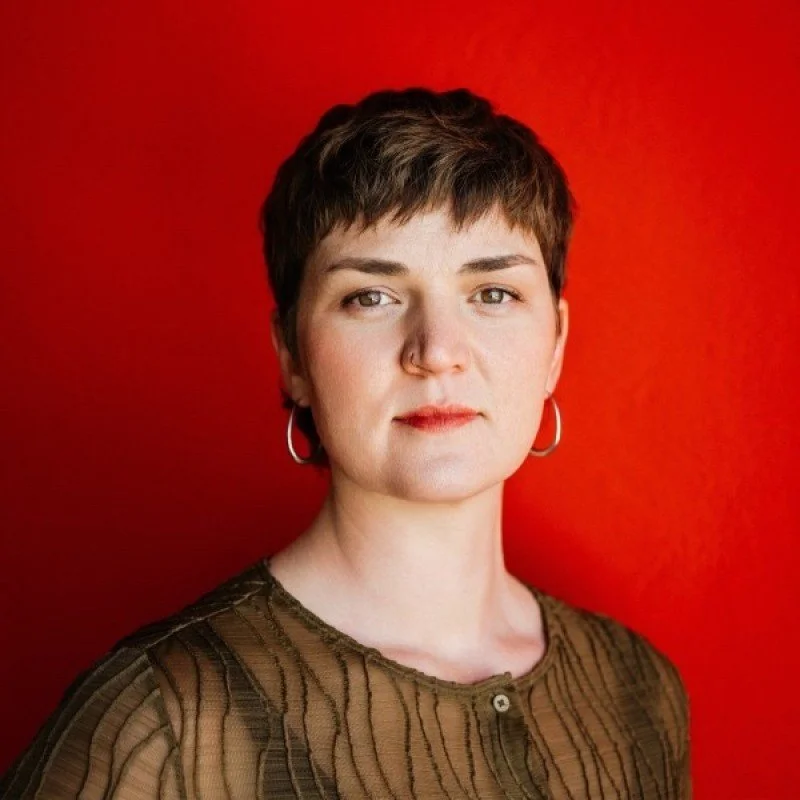Portrait of a woman with short brown hair, wearing hoop earrings and a brown patterned top, standing against a red background.