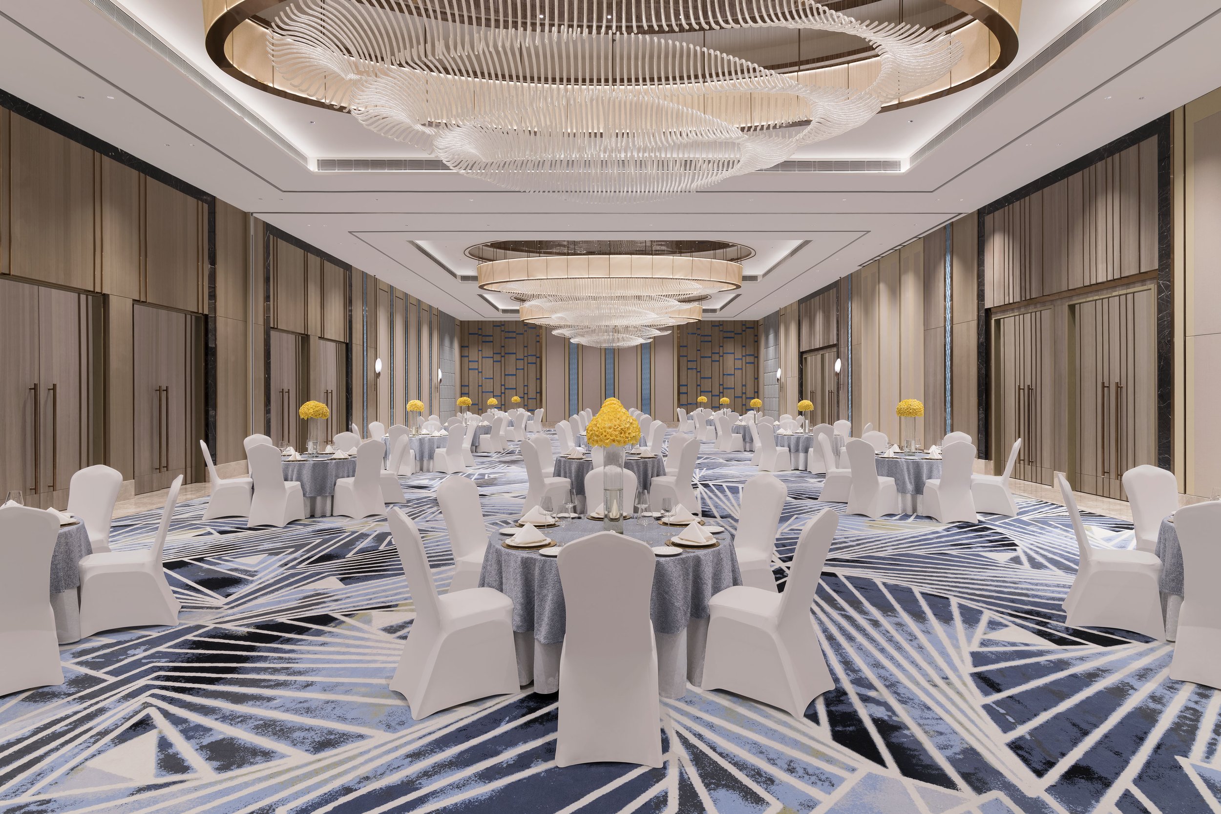 Elegant banquet hall with round tables draped in gray tablecloths, each set with white plates, napkins, and glassware. White chairs surround the tables, and yellow flower centerpieces adorn each. The room features a geometric patterned carpet, wood-paneled walls, and large, ornate chandeliers hanging from a white ceiling with recessed lighting.