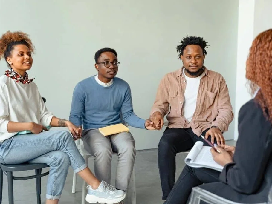 A group of four diverse people sitting in a circle during a support or counseling session, holding hands and engaging in a comforting or supportive activity.