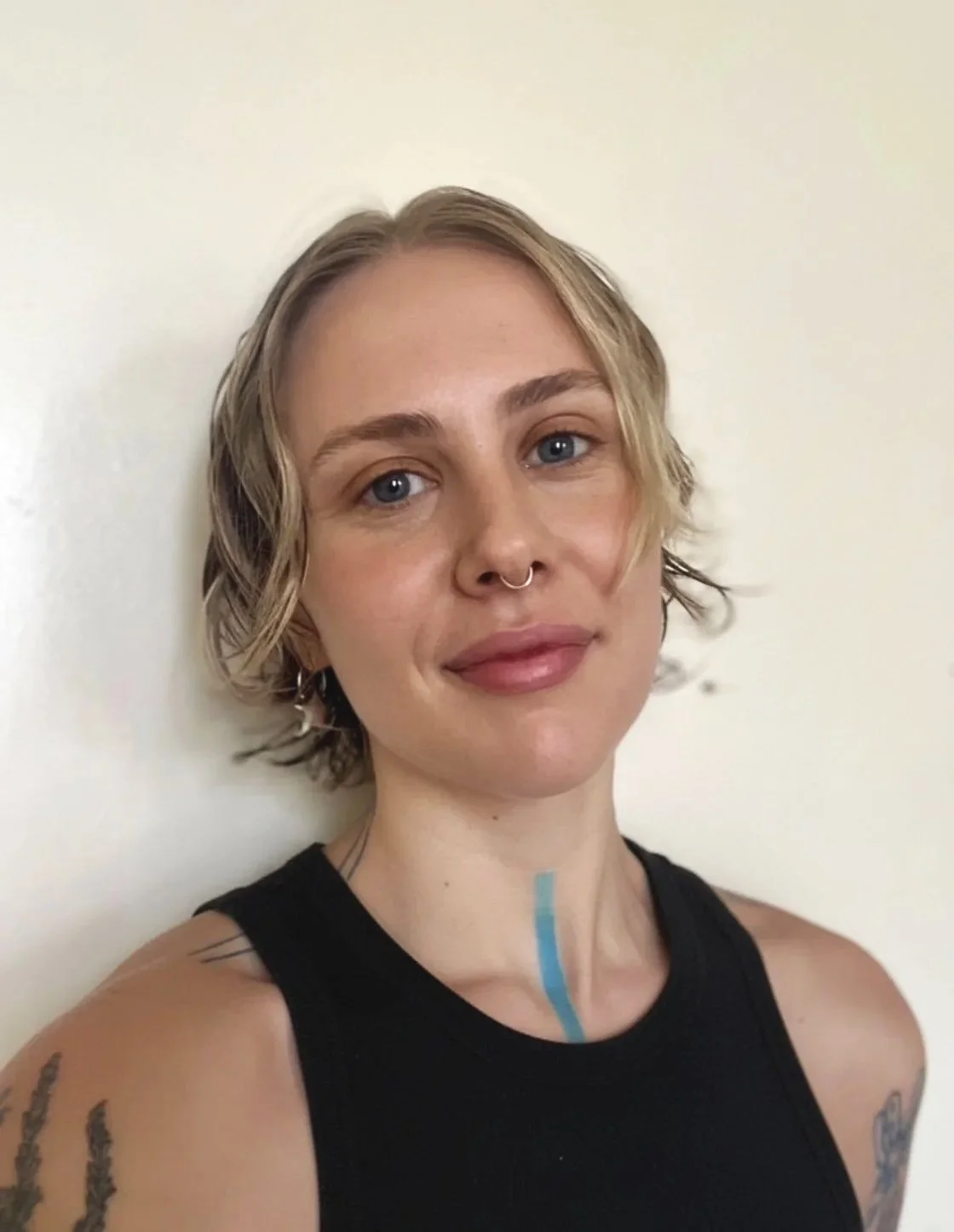 A woman with short blonde hair, blue eyes, wearing a black tank top, and a septum piercing, standing against a plain off-white wall.