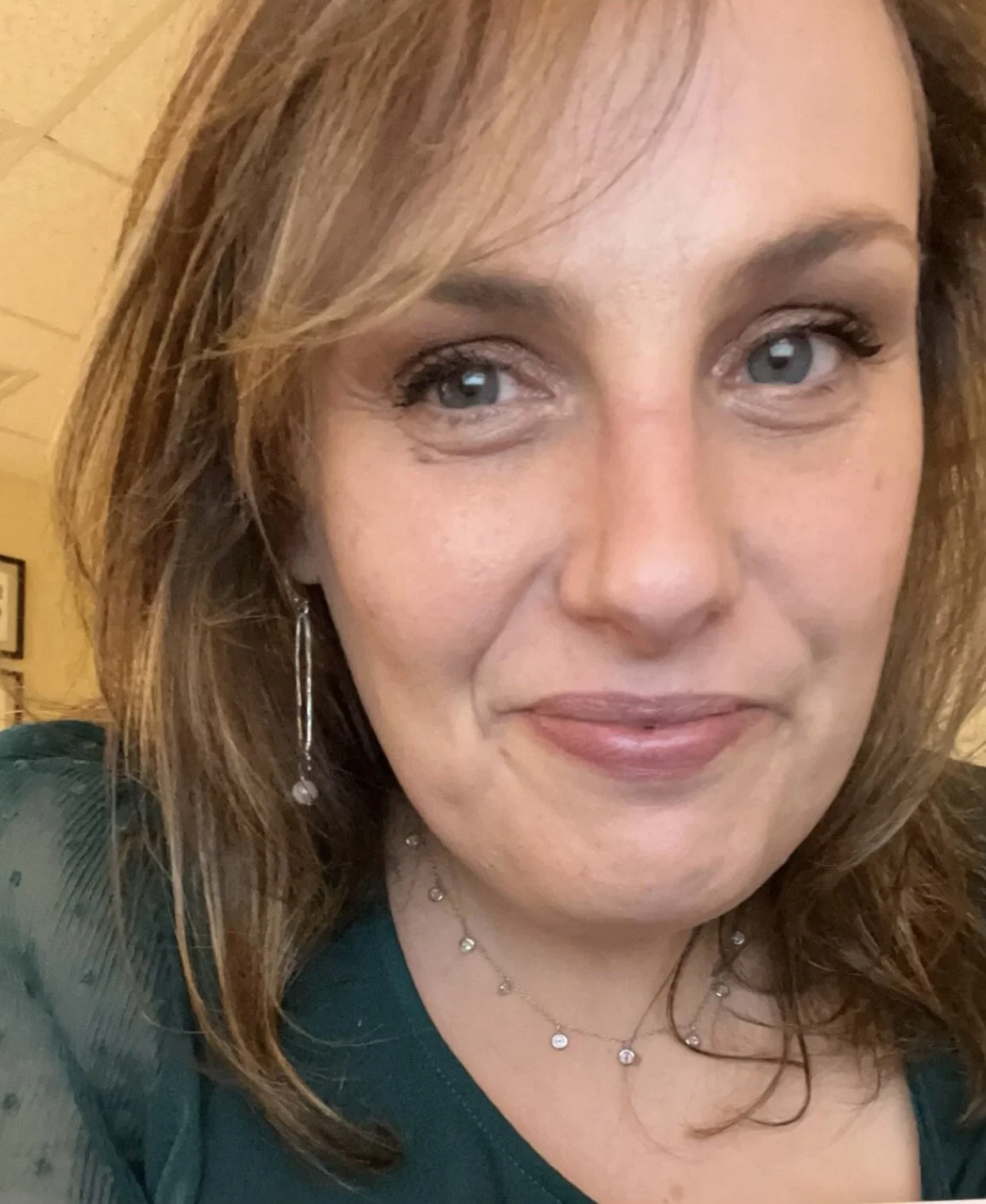 A woman with shoulder-length light brown hair and blue eyes smiling at the camera, wearing earrings and a necklace, in an indoor setting with a yellowish background.