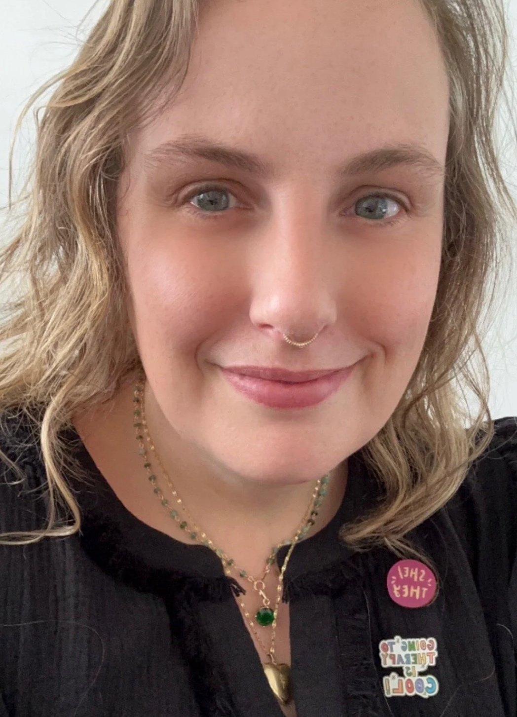 A smiling woman with blond wavy hair, wearing a septum ring, layered necklaces, and a black shirt with colorful buttons that say 'Cool! Don't Care!' and 'She/Her'.