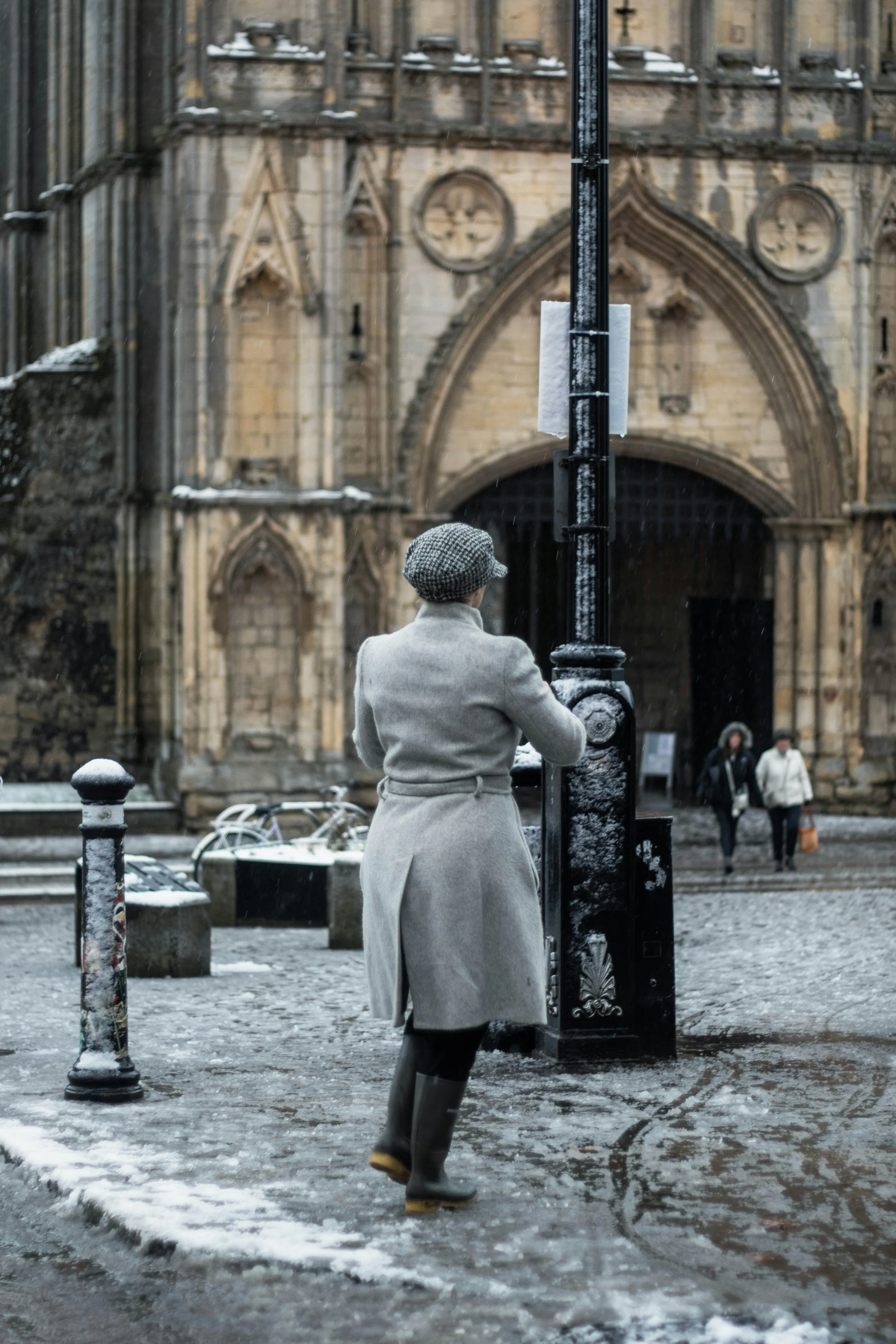 A woman in a gray coat, hat, and rain boots stands on a snowy cobblestone street, facing a lamppost in front of an old stone church.