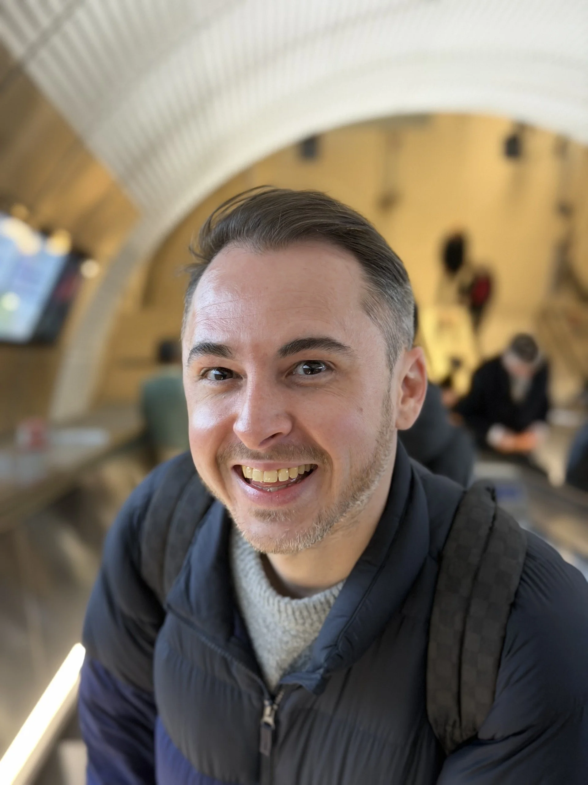 A man smiling at the camera inside a modern, curved architectural space with people in the background.