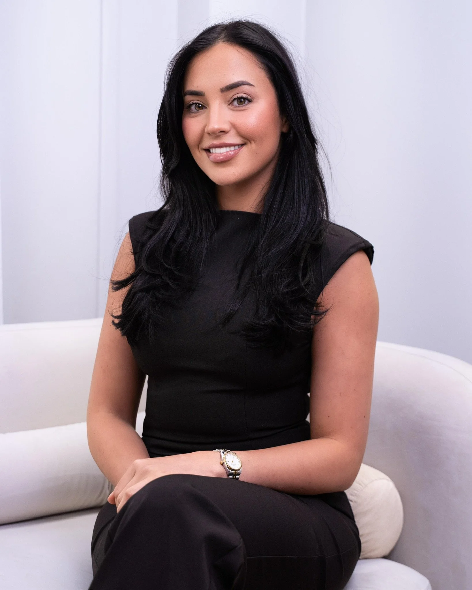 A woman with long black hair, wearing a black sleeveless dress, sitting on a white couch, smiling at the camera.
