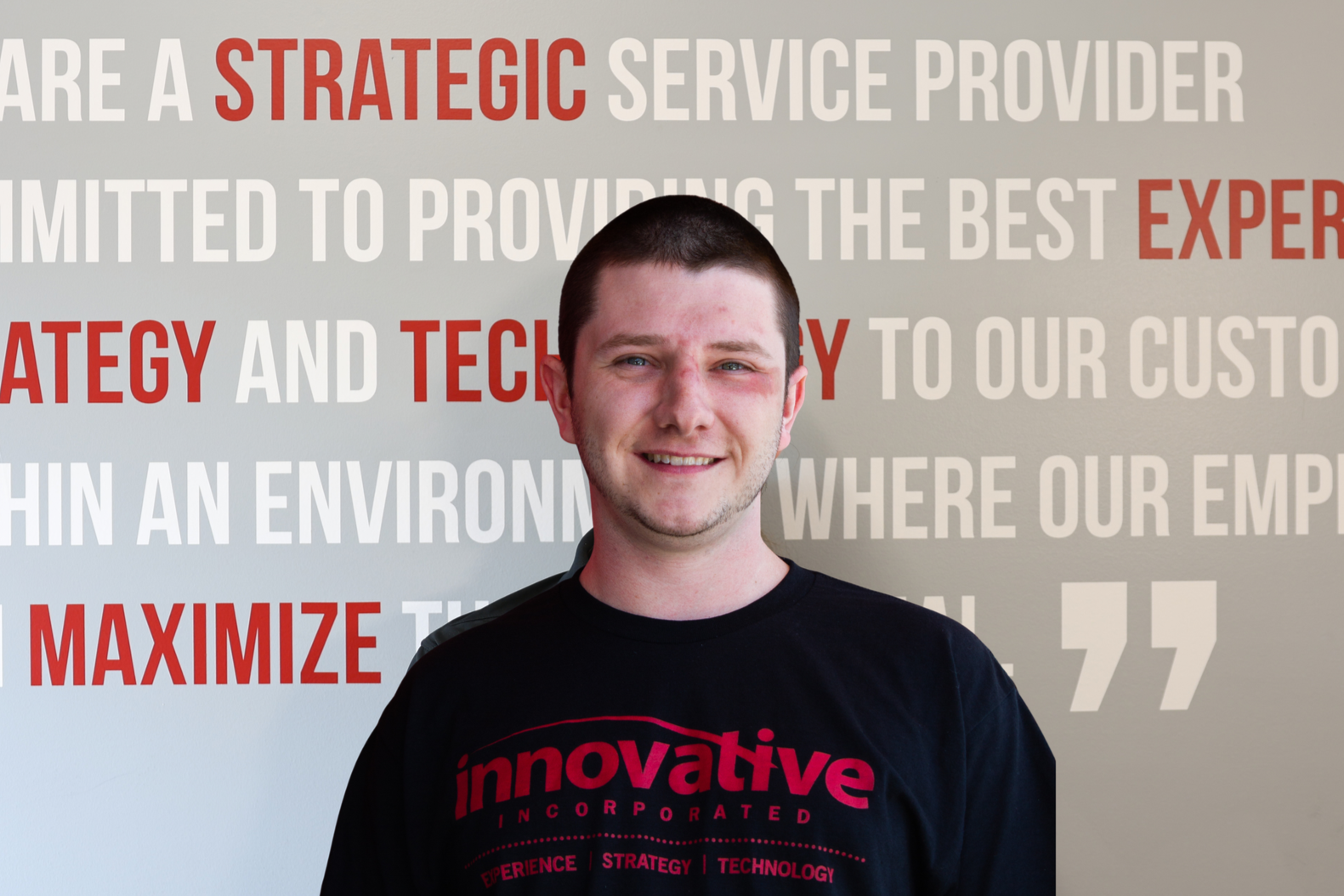 A young man with short dark hair, smiling, wearing a black T-shirt with red text, standing outside near a red and black building.