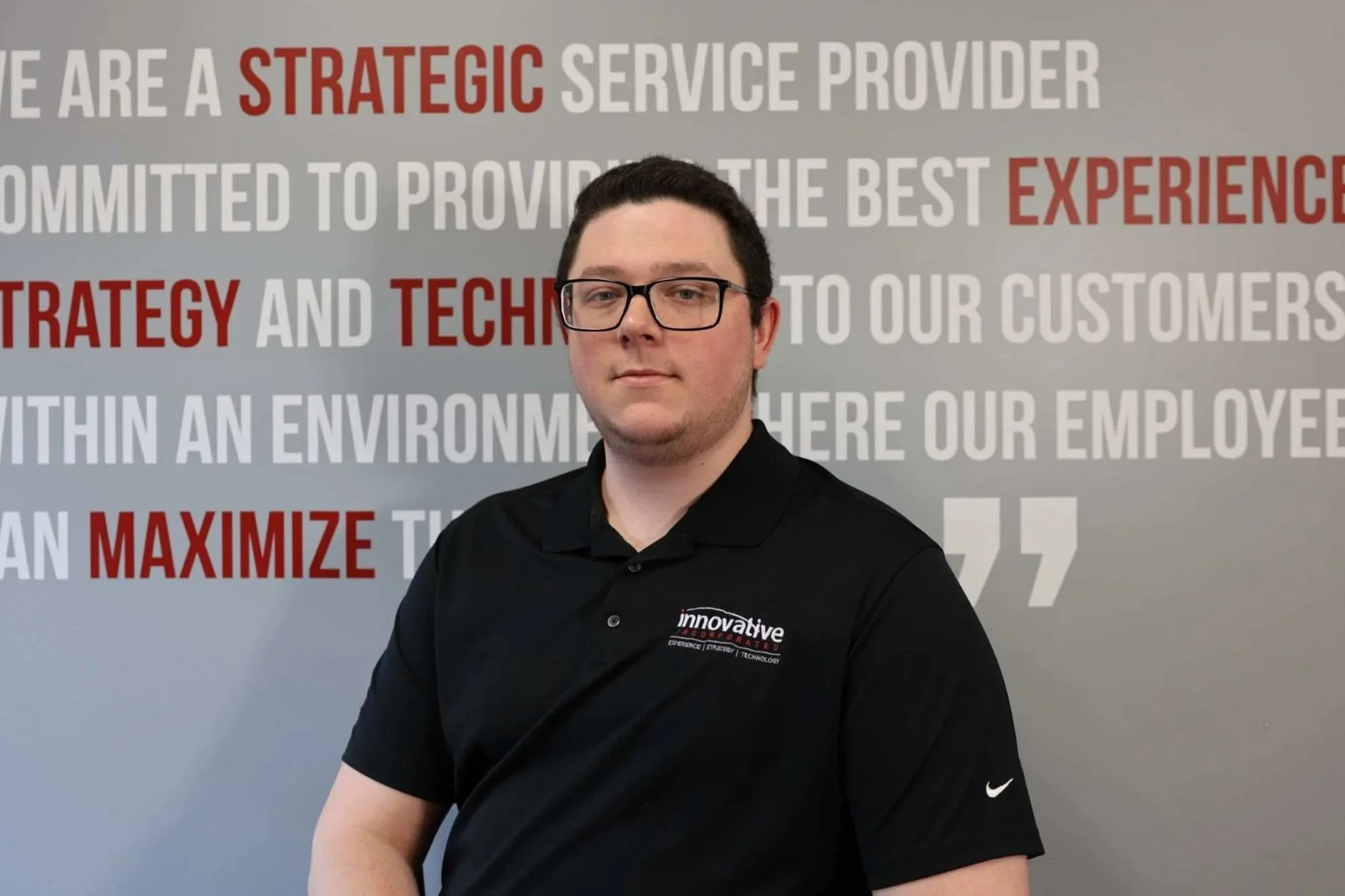 Young man with glasses wearing a black polo shirt with the logo 'Innovative Inc.' standing in front of a red metal fence with cars and trees in the background.