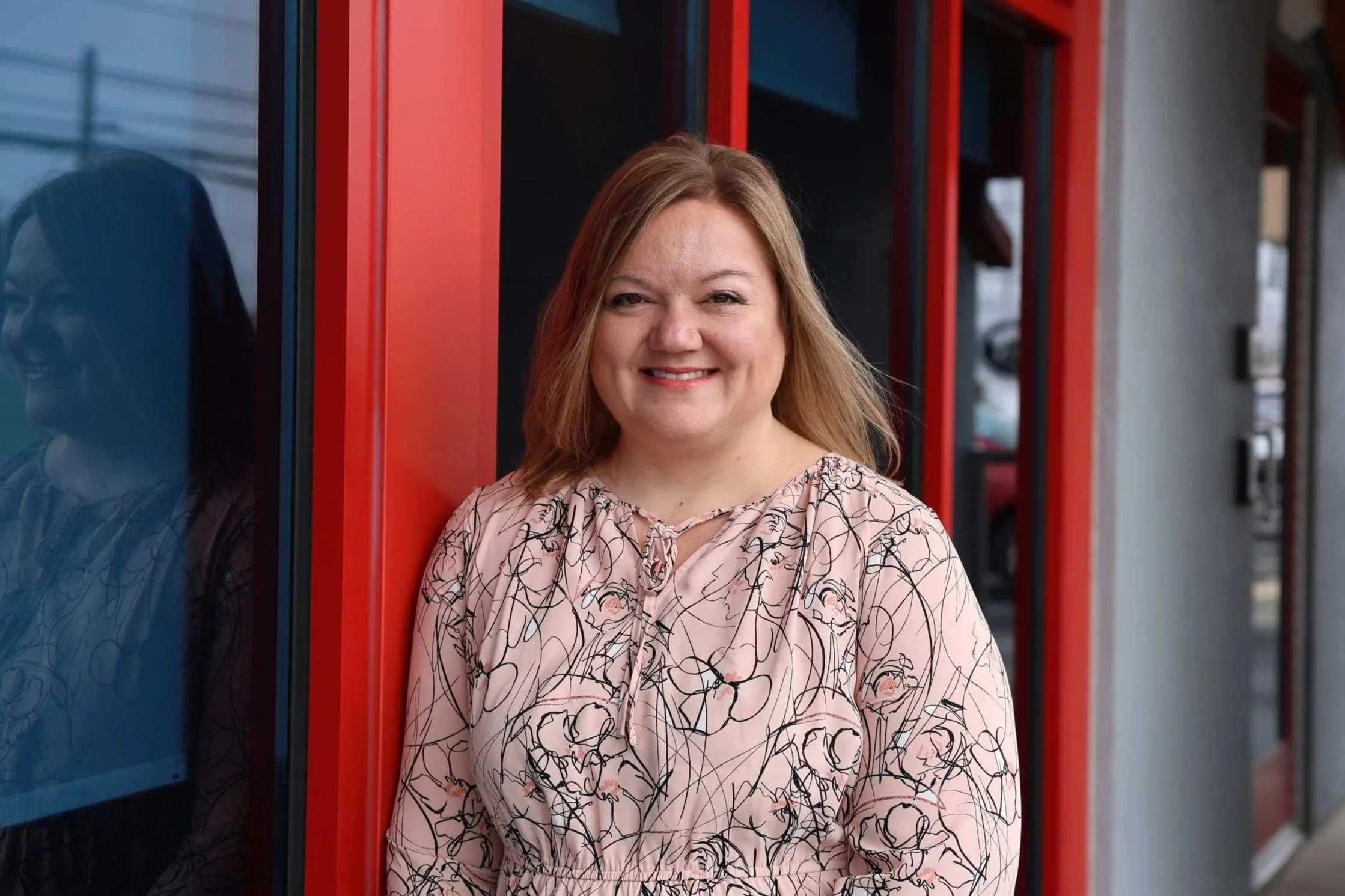 A smiling woman with shoulder-length brown hair, wearing a white top and navy blue cardigan, standing in front of a window with red framing.
