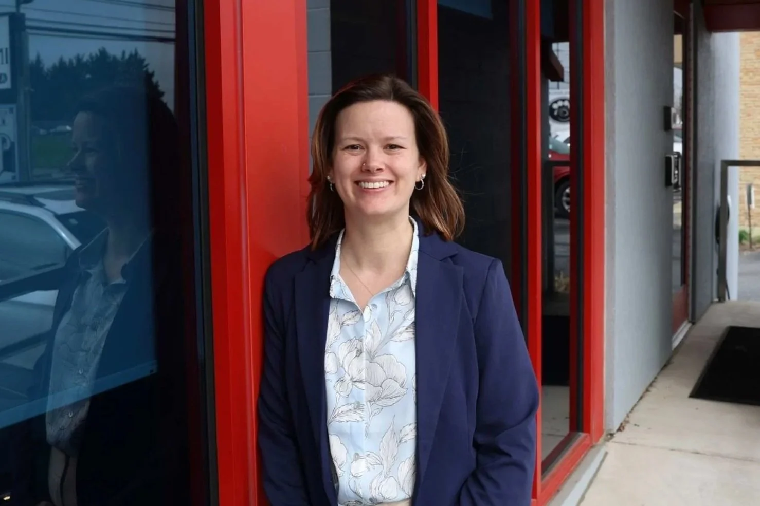 A young woman with shoulder-length brown hair smiling, standing next to a red wall with large windows.