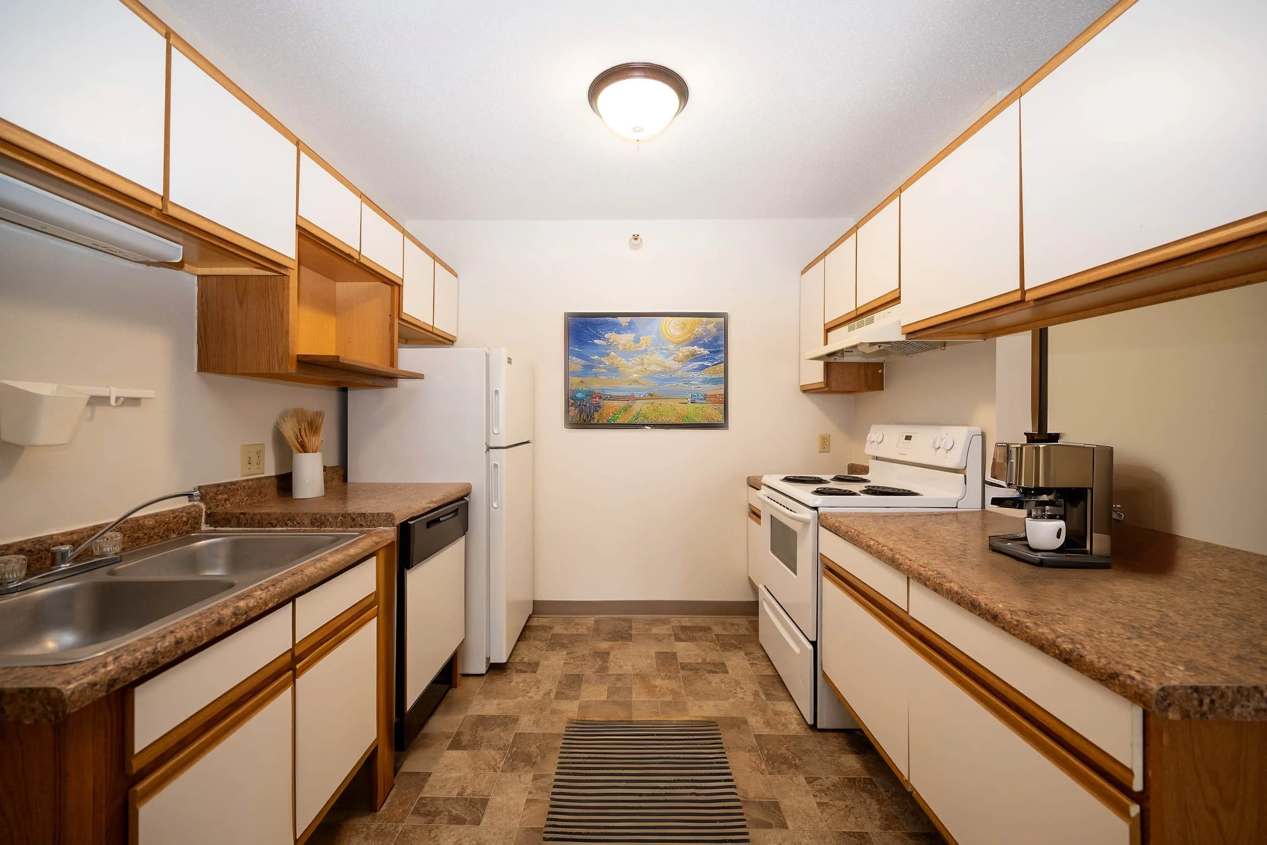 Galley kitchen in apartments in Rochester with white cabinets, brown countertops, striped rug, and a colorful painting on the back wall.