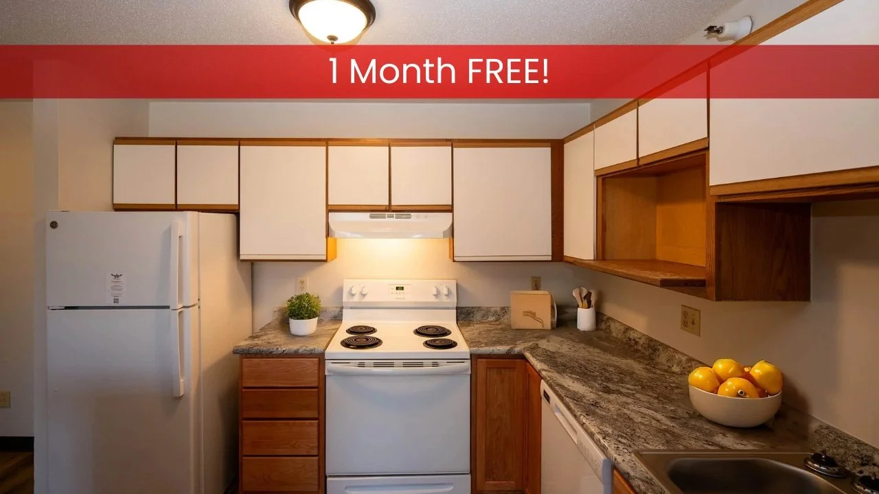 Small kitchen in apartments in Rochester with white cabinets, stove, fridge, utensils, a potted plant, and a bowl of apples.