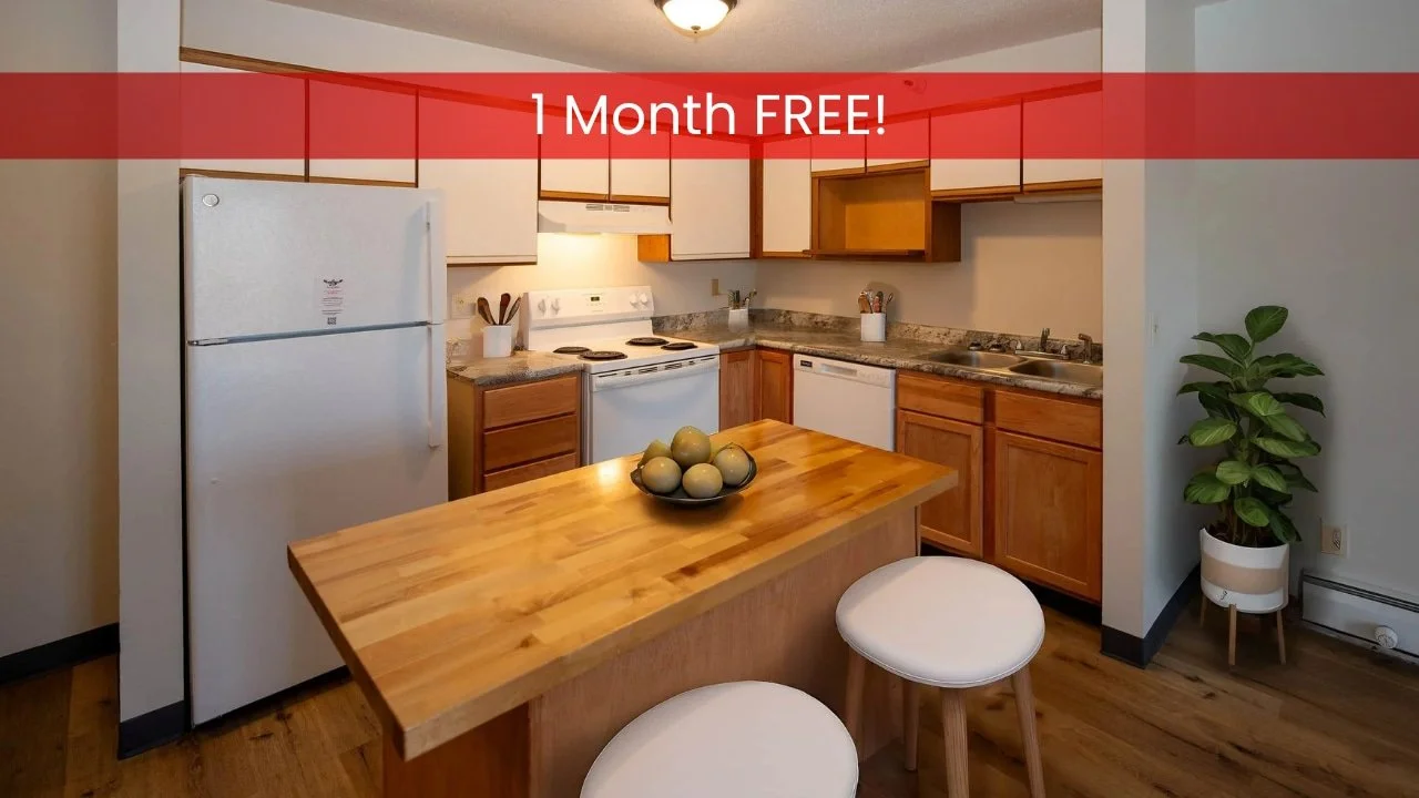 Modern kitchen in apartments in Rochester features white appliances, wood cabinets, an island with fruit, stools, and a potted plant.