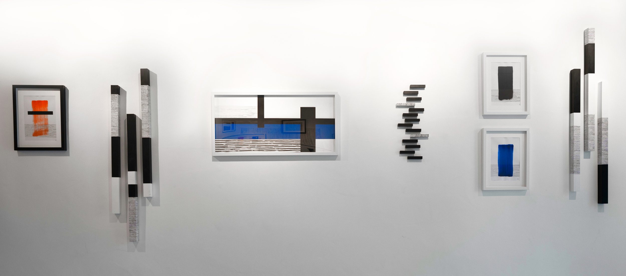 Abstract mixed-media art installation on a white gallery wall, including framed paintings with bold colors, vertical and staggered rectangular sculptures, and a rectangular window with a reflective surface showing blue and black geometric patterns.