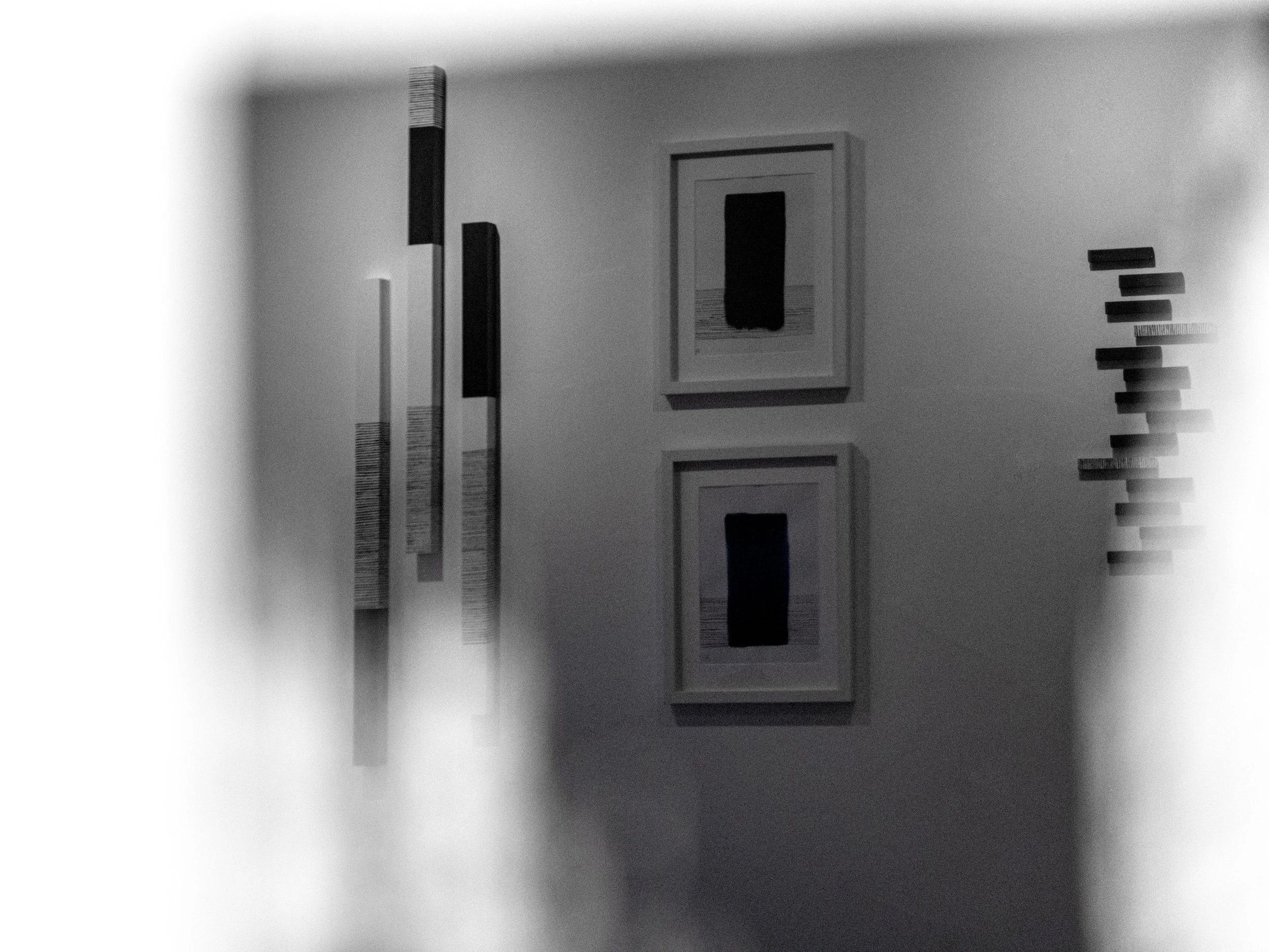Black and white photo of framed artwork, vertical rectangular, on wall with horizontal rectangular artwork to the right, also framed, and several small abstract pieces arranged vertically.