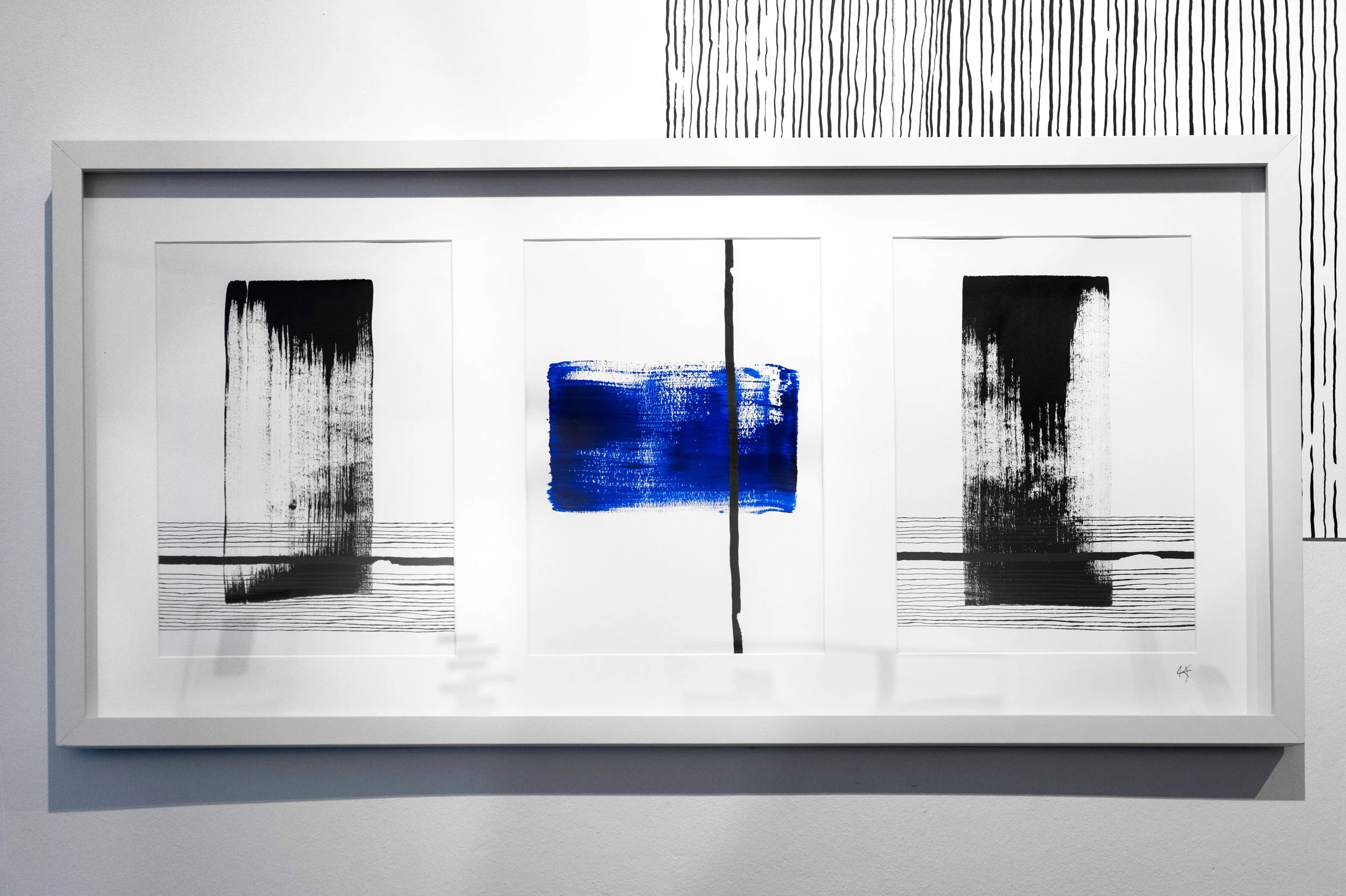 A white framed triptych art piece with black and blue abstract brushstrokes, set against a white wall with black line patterns.