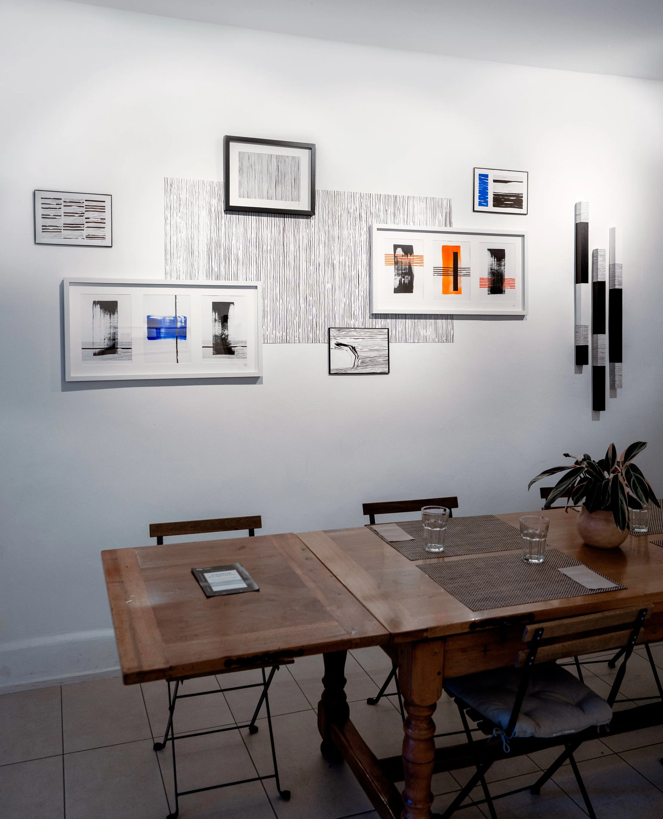 Gallery wall with abstract artwork, wooden table with glasses of water and potted plant, and wooden chairs in a modern dining area.