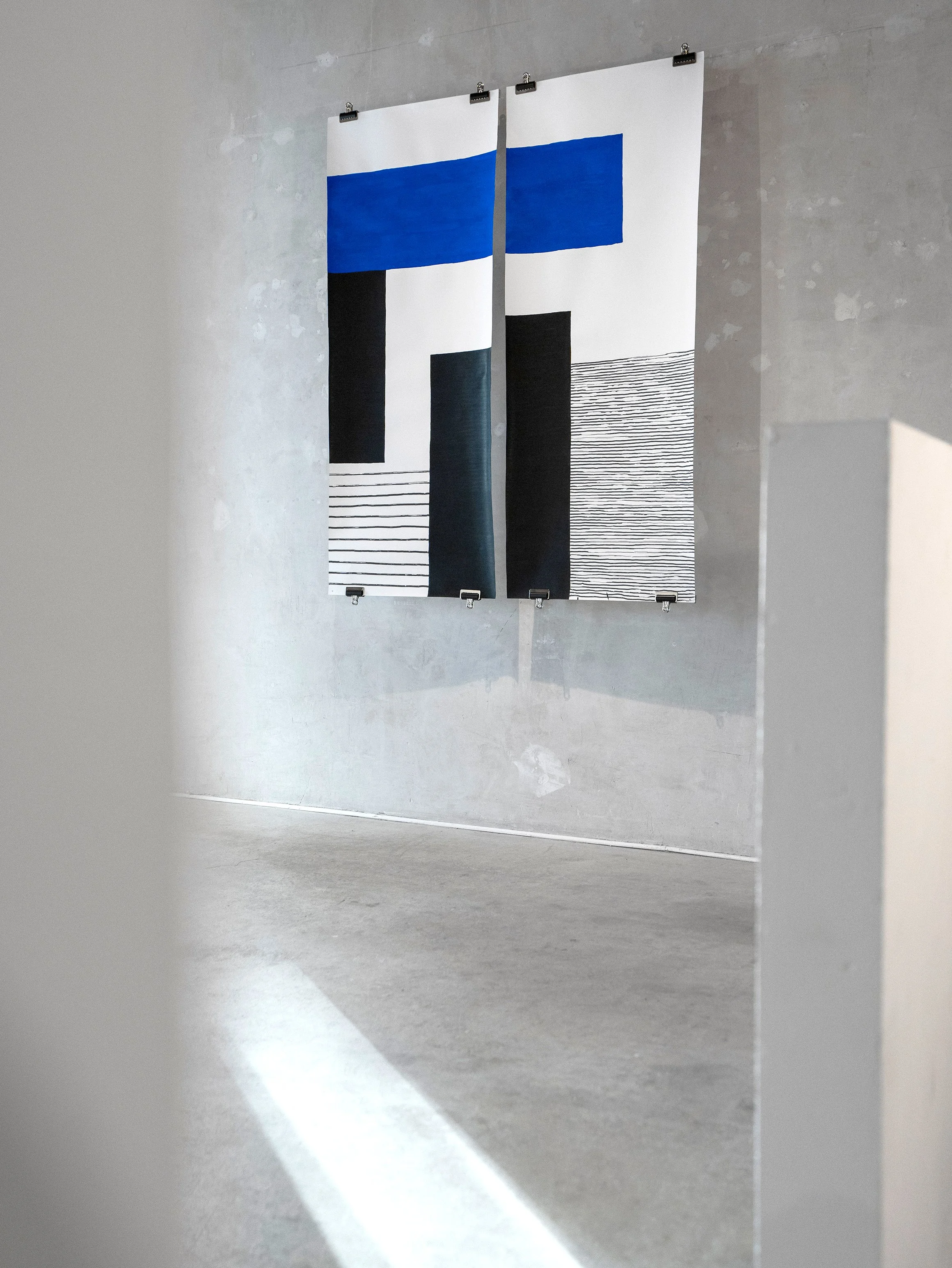 Two modern abstract fabric wall art pieces with geometric shapes and horizontal lines in black, white, and blue hanging on a concrete wall.