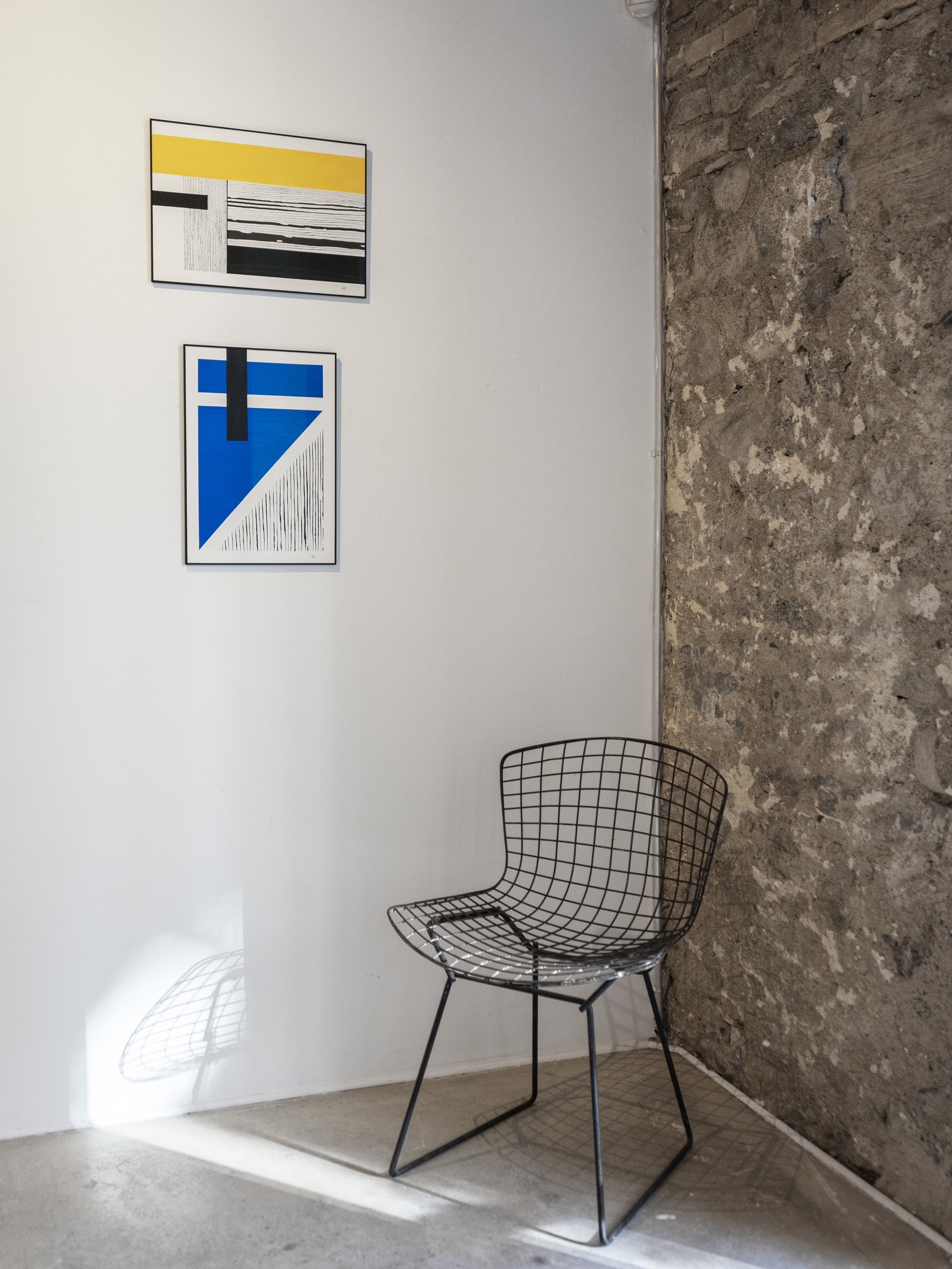 Empty wire chair in front of a white wall with two modern abstract paintings, one with yellow, black, and white stripes, and the other blue with black accents, on a polished concrete floor next to a rough, exposed stone wall.