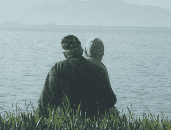 A man and a young boy, seen from behind, sitting close together at the edge of calm water, looking out across it.