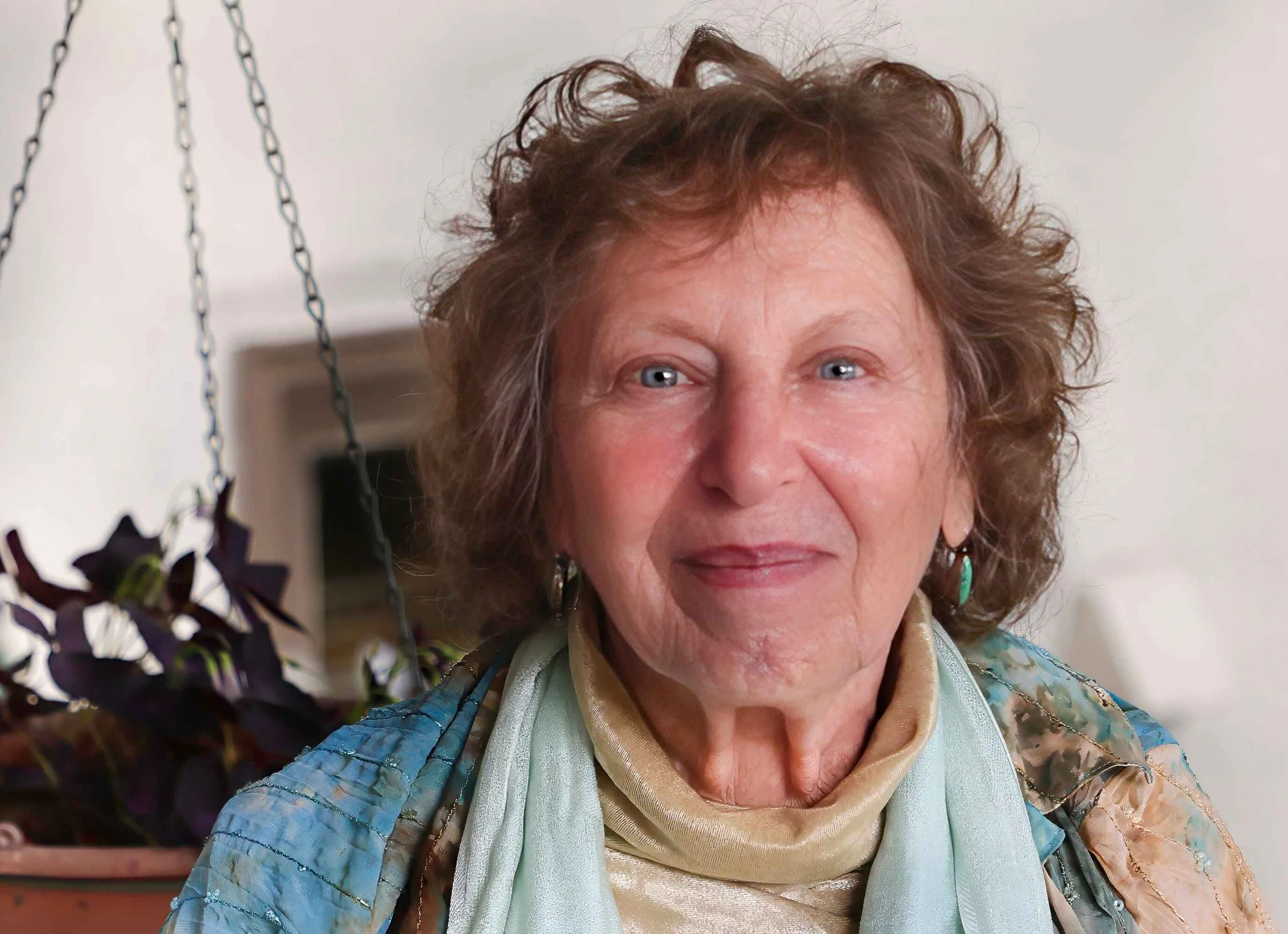 Author photo of Naomi Rose, a woman with curly auburn hair and blue eyes, wearing a soft blue scarf and layered clothing, smiling warmly, with a hanging plant visible in the background.