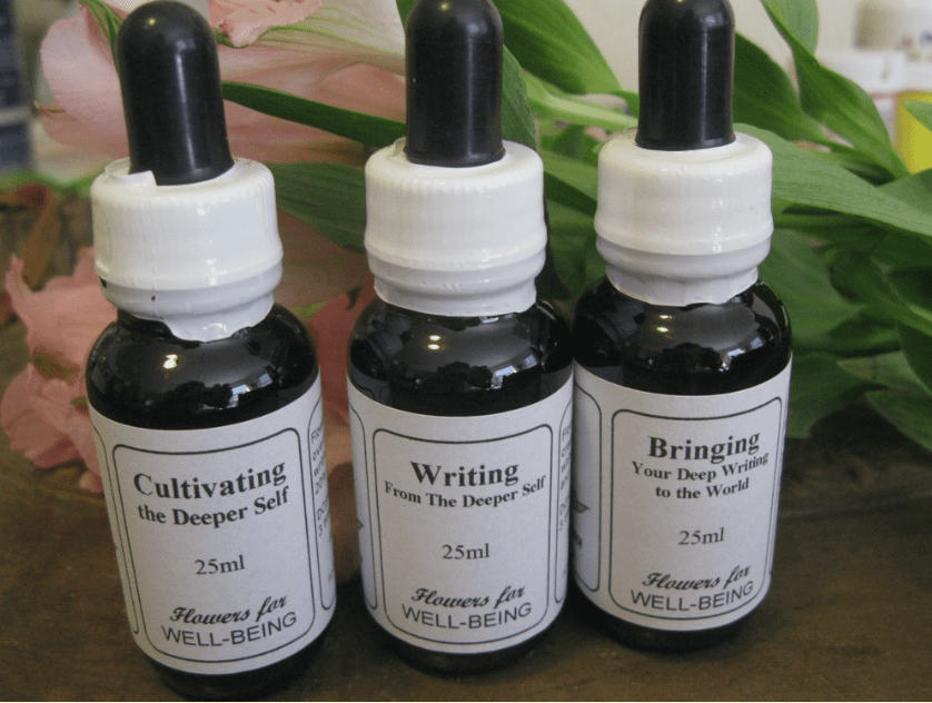 Three small black bottles with white labels and dropper caps, labeled "Cultivating the Deeper Self," "Writing From The Deeper Self," and "Bringing Your Deep Writing to the World," placed on a wooden surface with pink and green decorative leaves in the background.