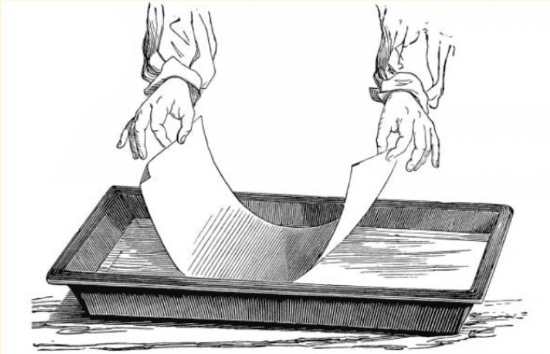 A vintage line drawing of two hands placing a photographic print into a developing tray