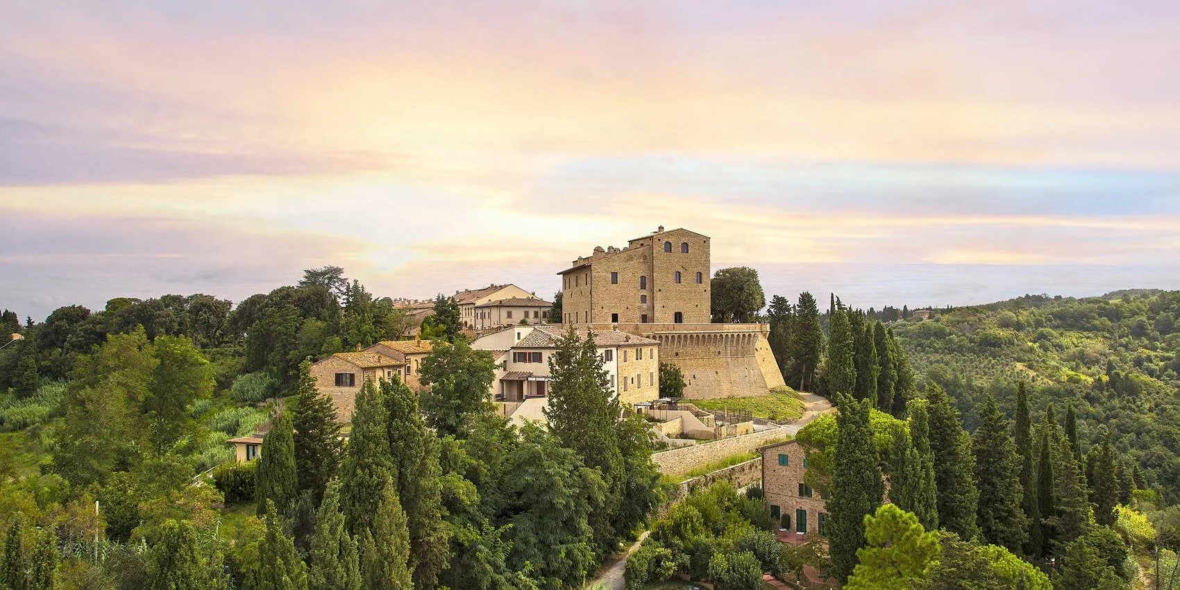 A scenic landscape featuring historic stone buildings perched on a hilltop, surrounded by lush green trees, under a pastel-colored sky at sunset in a rural area.