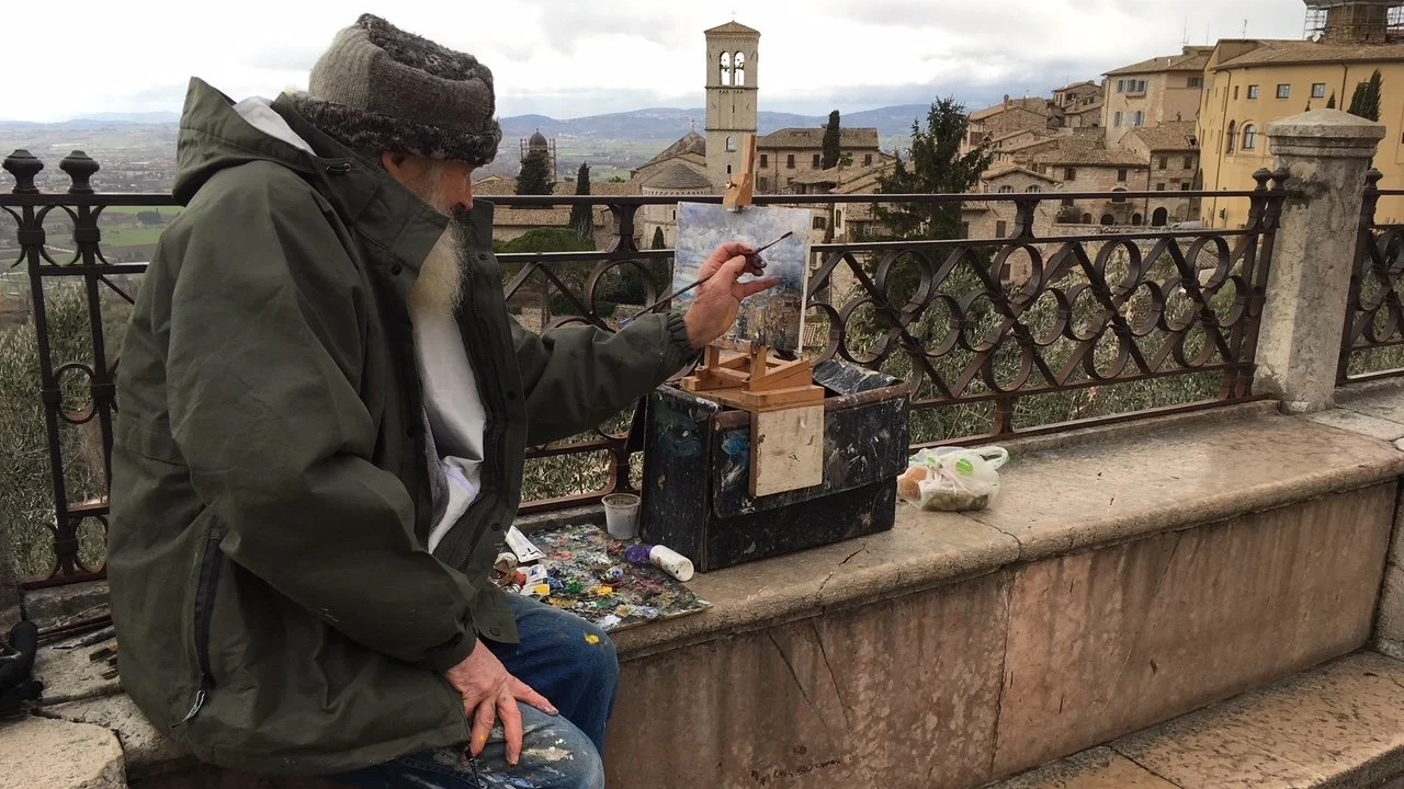 A man painting on a small canvas set up on a portable easel on a stone ledge outdoors, with a cityscape and hills in the background