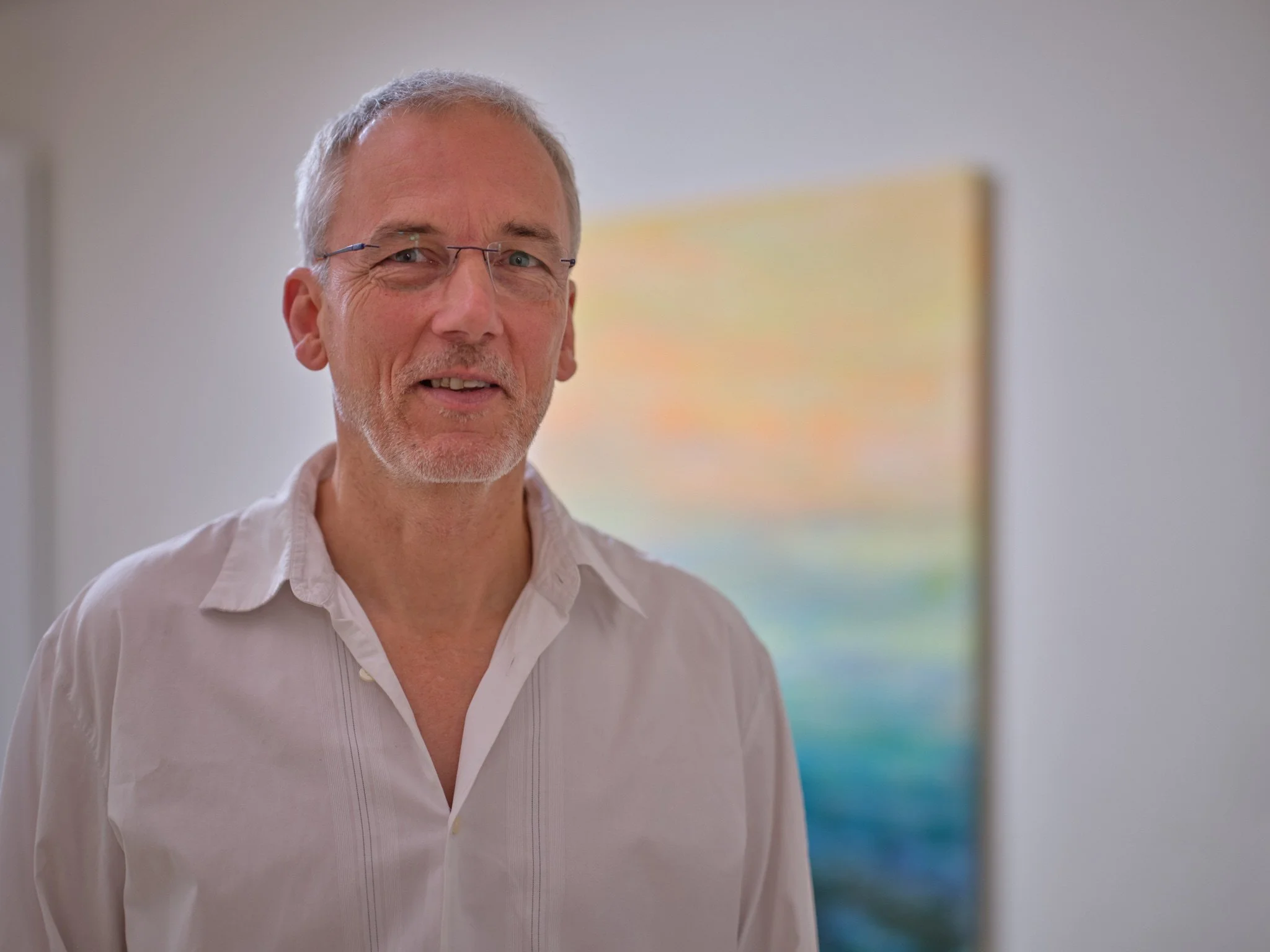 Andreas Brieschke mit Brille, weißem Hemd und grauem kurzen Haar, die im Vordergrund steht, mit einem bunten Kunstwerk im Hintergrund.