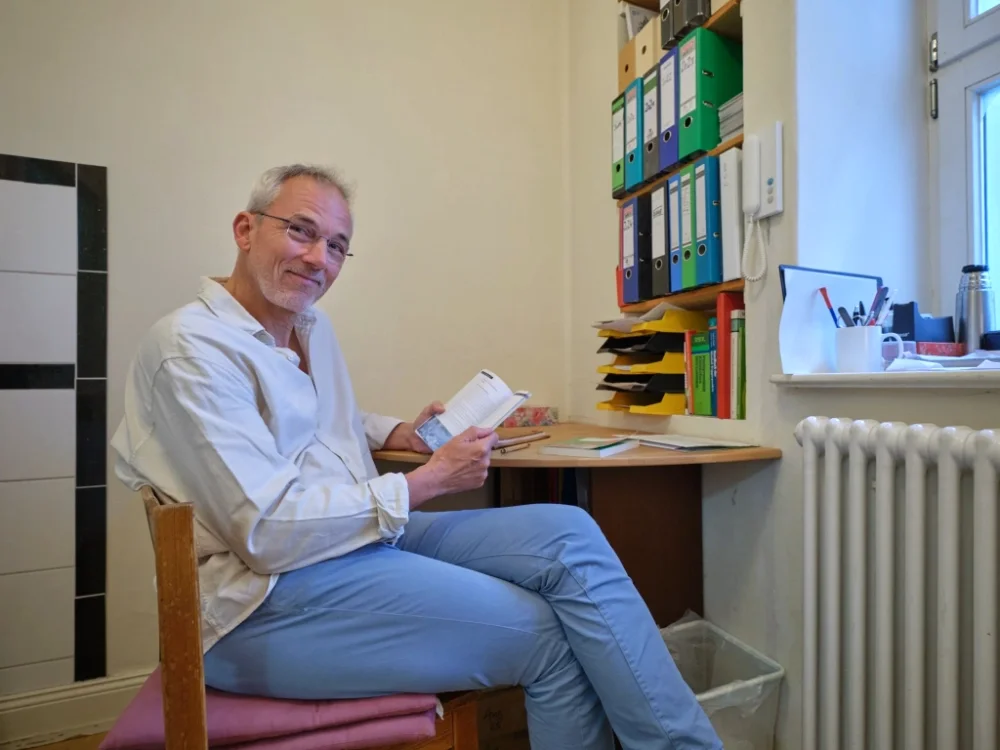 Ein älterer Mann sitzt in einem kleinen Büro, liest in einem Buch und lächelt. Im Hintergrund sind Aktenordner, Papiere und Stifte auf einem Regal und Schreibtisch. Es gibt ein Fenster mit Blick nach draußen.