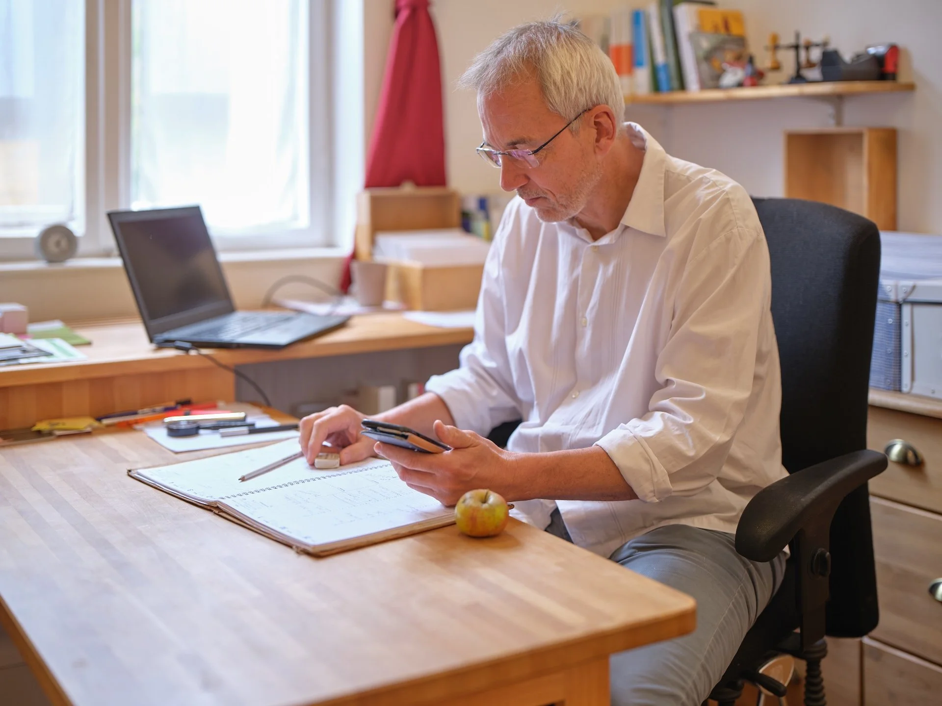 Andreas Brieschke  sitzt an einem Holztisch, schaut auf sein Smartphone, mit einem Notizblock und einem Apfel auf dem Tisch