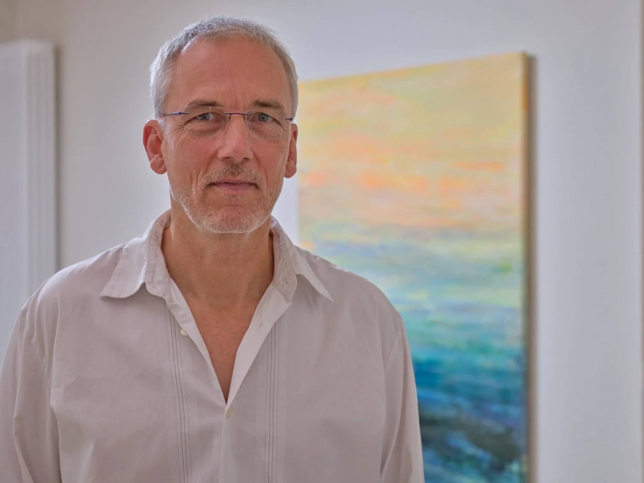 Ein älterer Mann mit kurzen grauen Haaren, Brille und weißem Hemd steht vor einer bunten, abstrakten Malerei an der Wand.