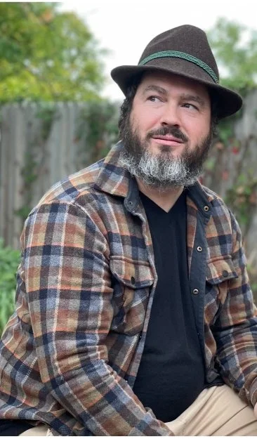 Man with a beard wearing a brown, green, and blue plaid shirt, black T-shirt, and a gray hat with a green band, sitting outdoors with a wooden fence and trees in the background.