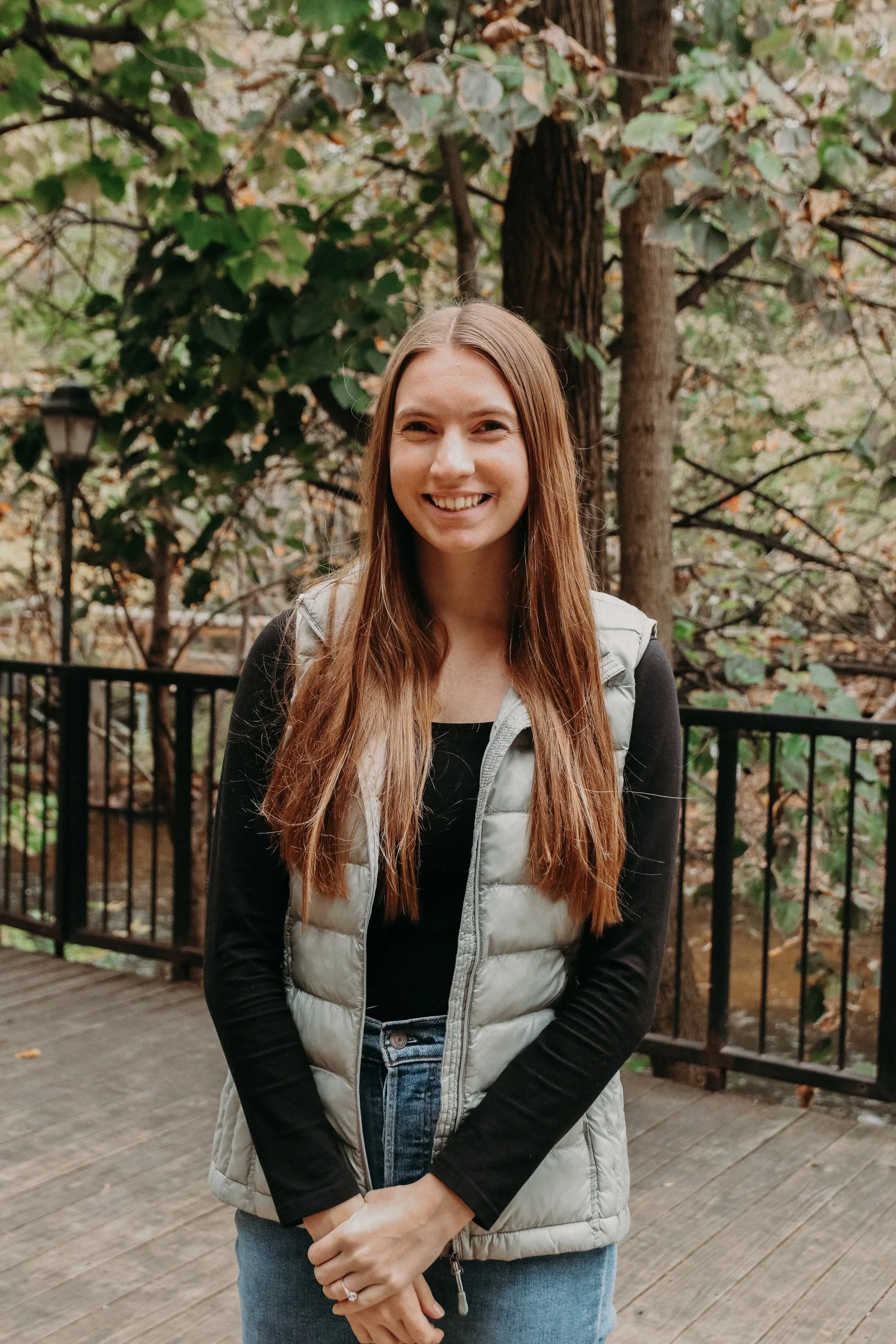 A young woman with long brown hair, smiling, standing outdoors on a wooden deck with a black railing, surrounded by trees and foliage.