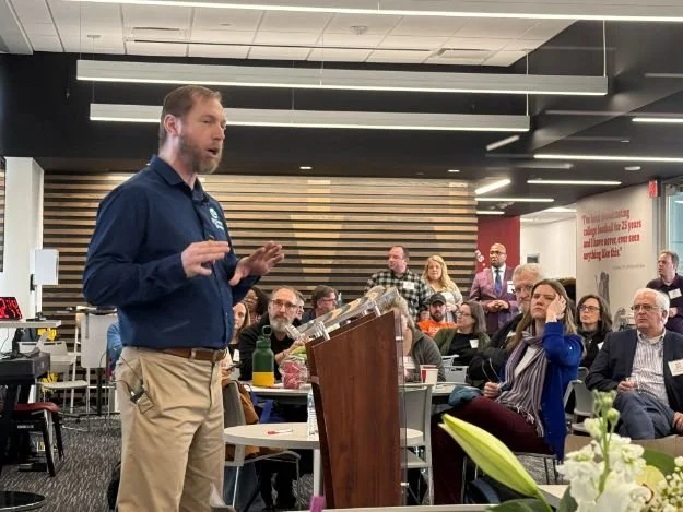 A man speaking at a podium during a conference or seminar, with an audience seated and listening attentively in a modern room with striped wood wall and ceiling lights.