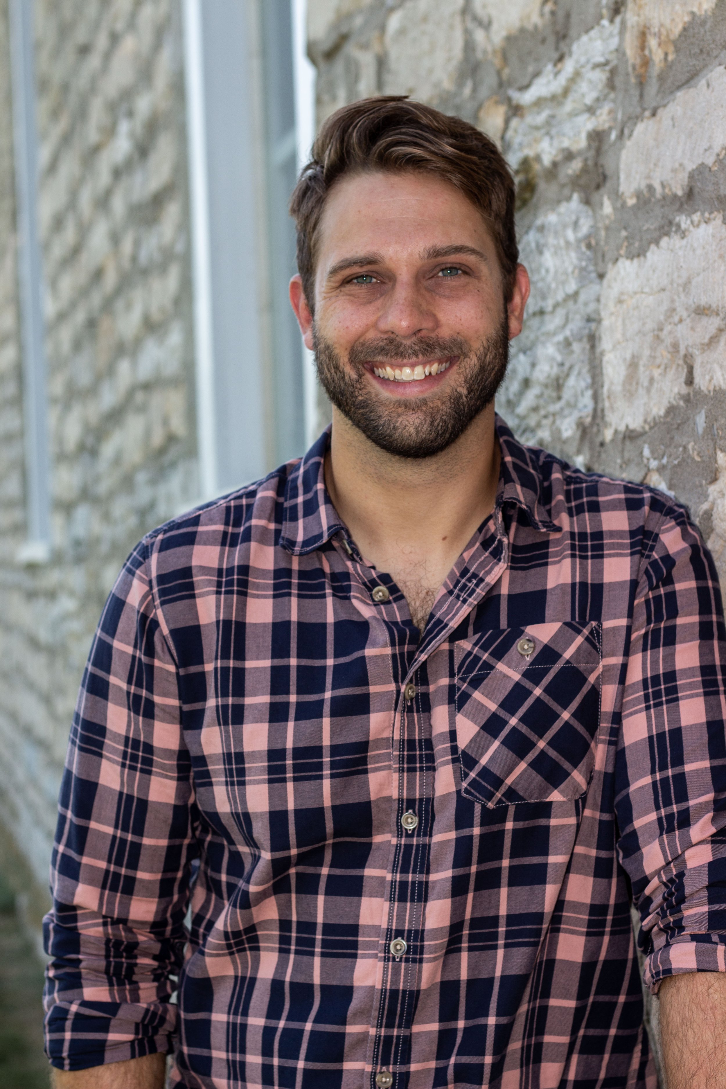 A man with a beard smiling outdoors near a stone wall, wearing a pink and navy plaid shirt.
