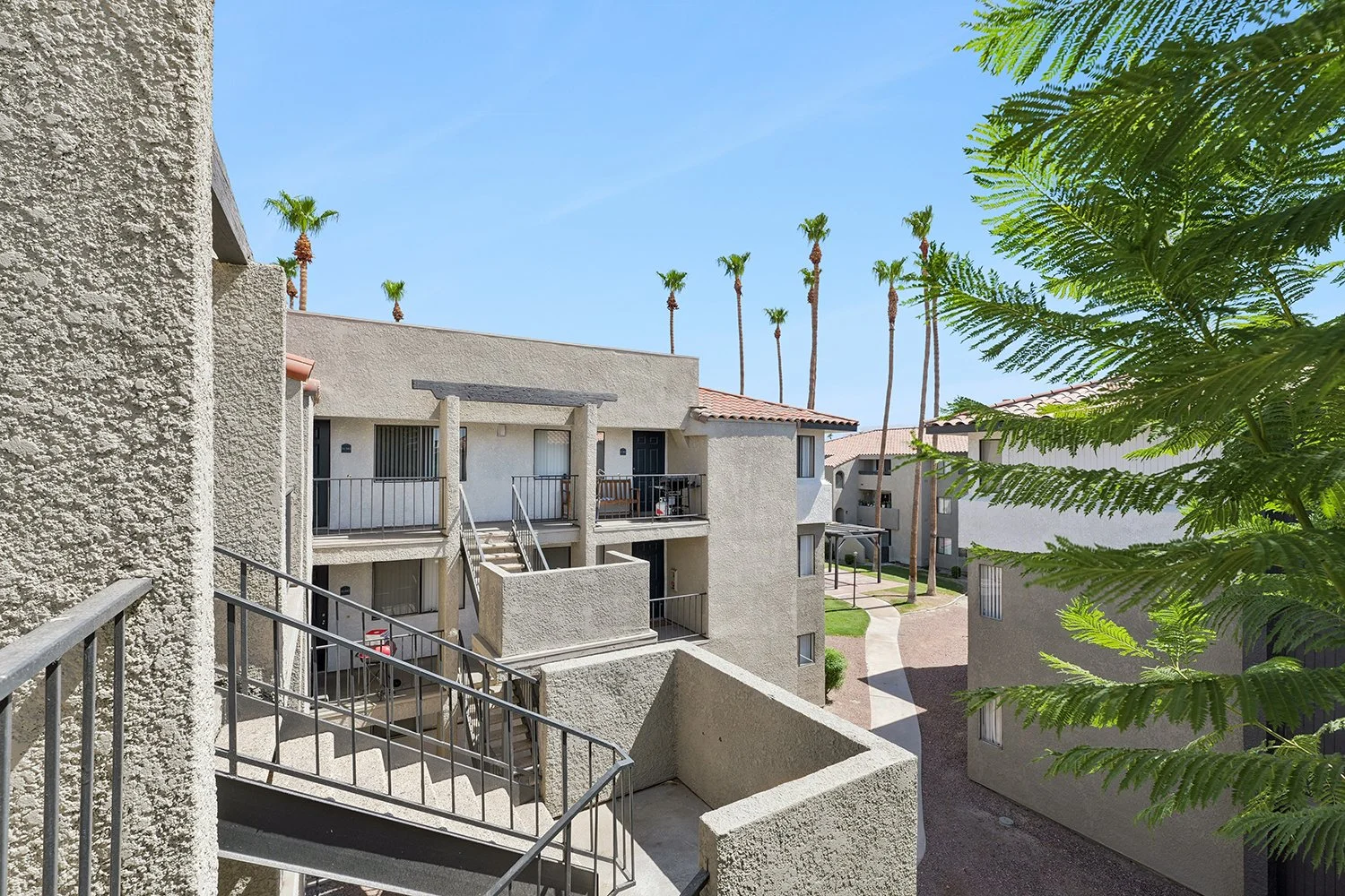 This image shows an elevated view of the inner courtyard, highlighting multi-story buildings with outdoor staircases and black metal railings. A concrete walkway winds through the landscaped grounds in the background. 