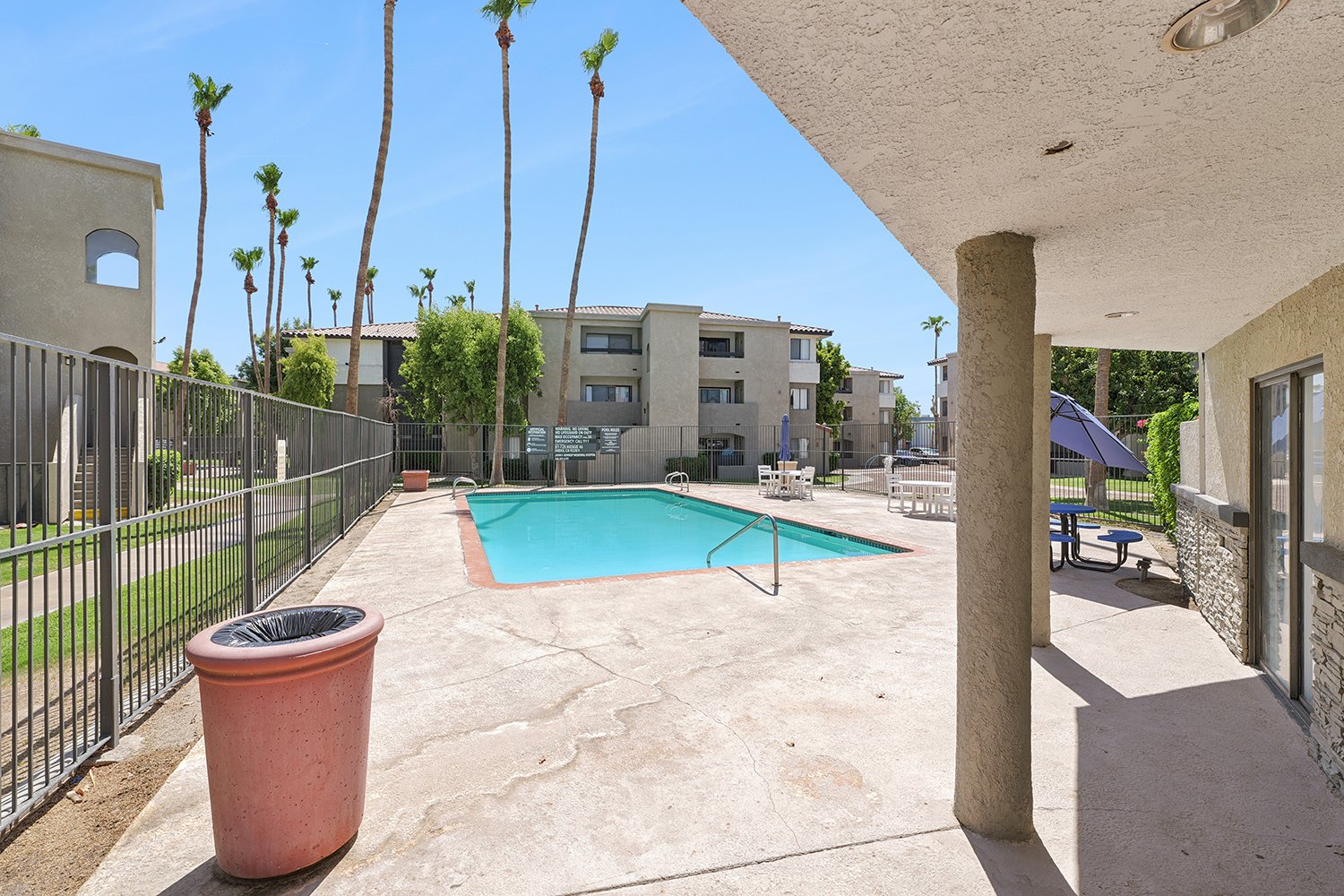 An image taken under a concrete awning looking out at a rectangular swimming pool and several outdoor tables and umbrellas. 