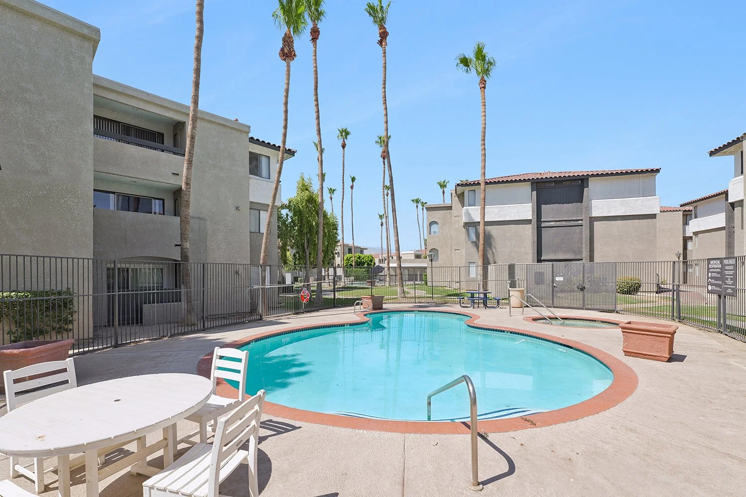 An image that captures the apartment community's fenced-in swimming pool area, featuring a curved blue pool and a small, adjacent hot tub with seating and a table. 